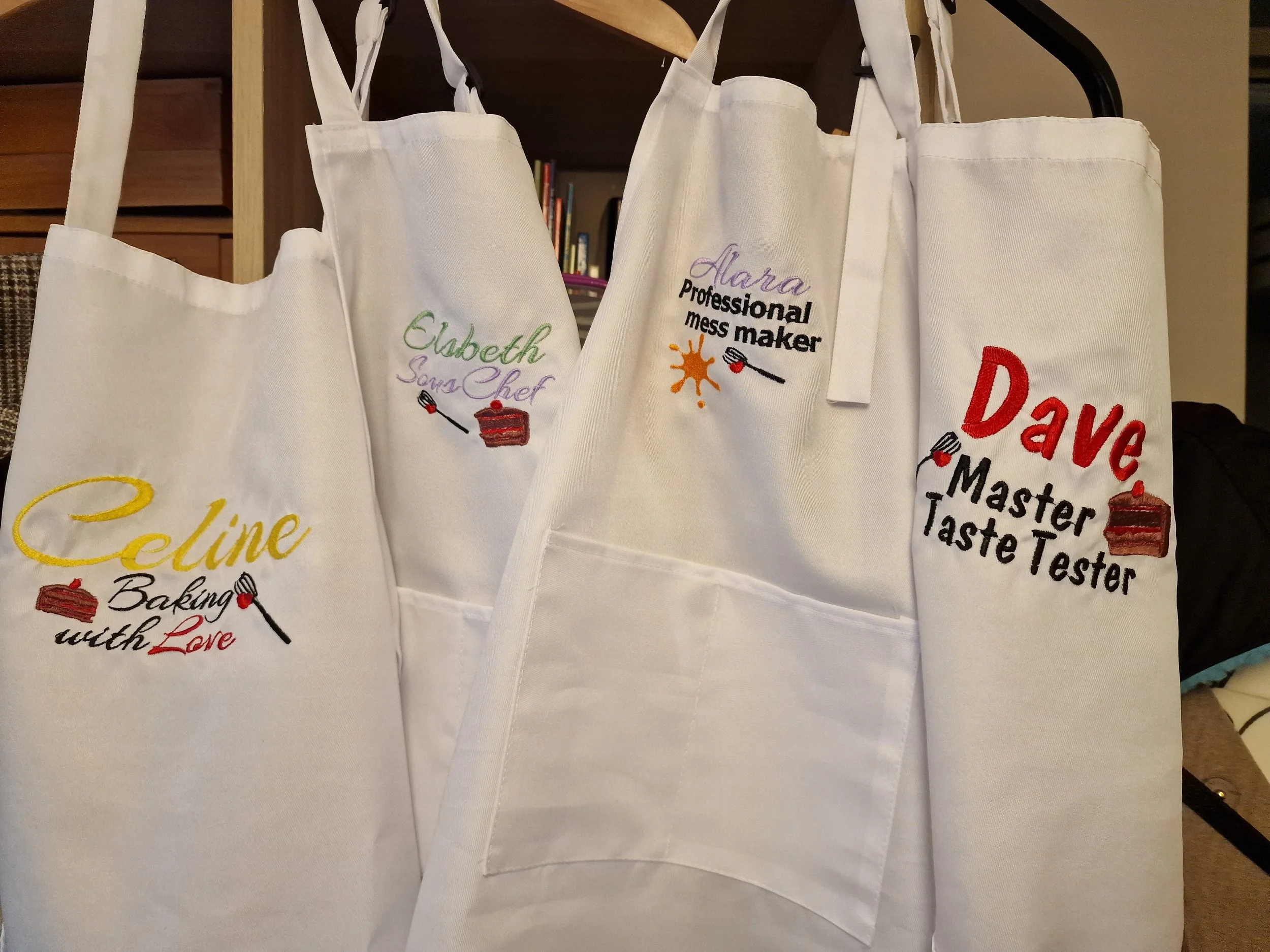 White aprons embroidered with colorful names and titles, including 'Celine Baking with Love', 'Elizabeth Sous Chef', 'Alara Professional Mess Maker', and 'Dave Master Taste Tester', each featuring a small cake and whisk graphic