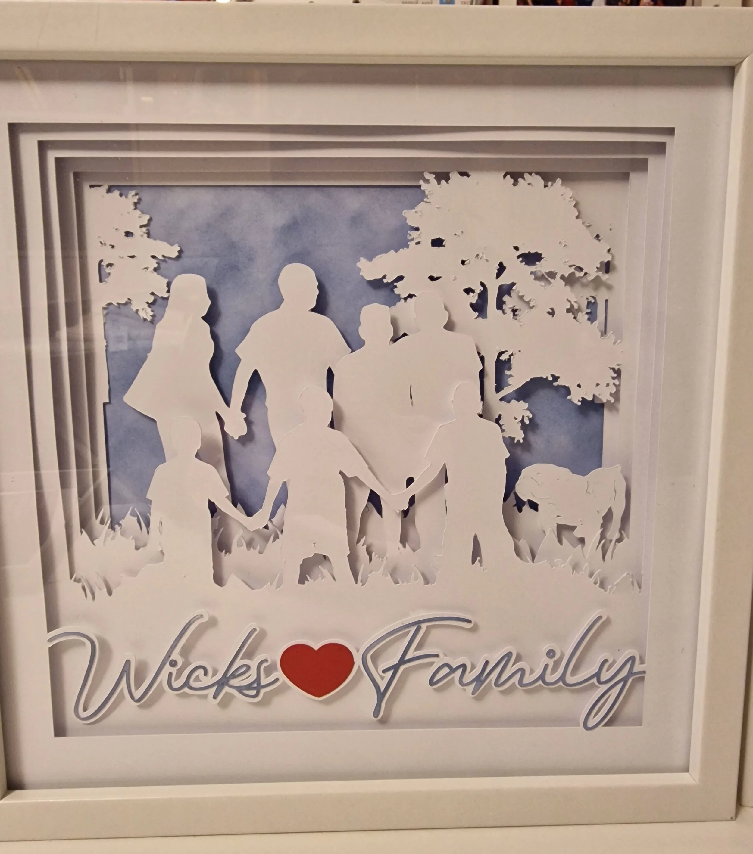 Paper cutout silhouette of a family holding hands outdoors with trees and grass, and the words 'Wicks Family' with a red heart in place of the dot in the letter 'i'.
