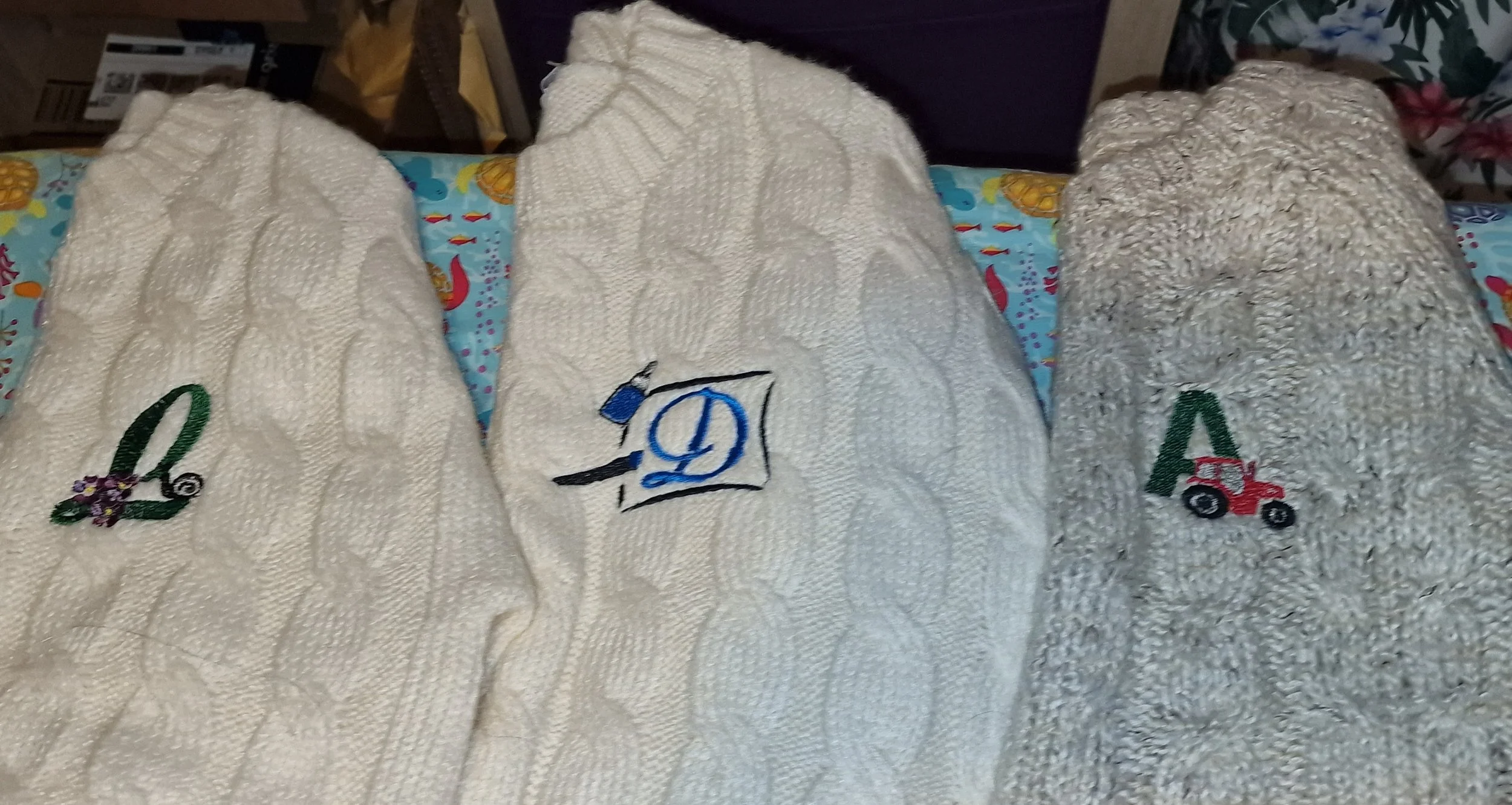 Three embroidered towels with different designs, placed on a colorful tablecloth with a marine theme. The towels have a cable knit pattern. The left towel features a green letter 'L' with flowers, the middle towel has a blue letter 'D' with a sewing