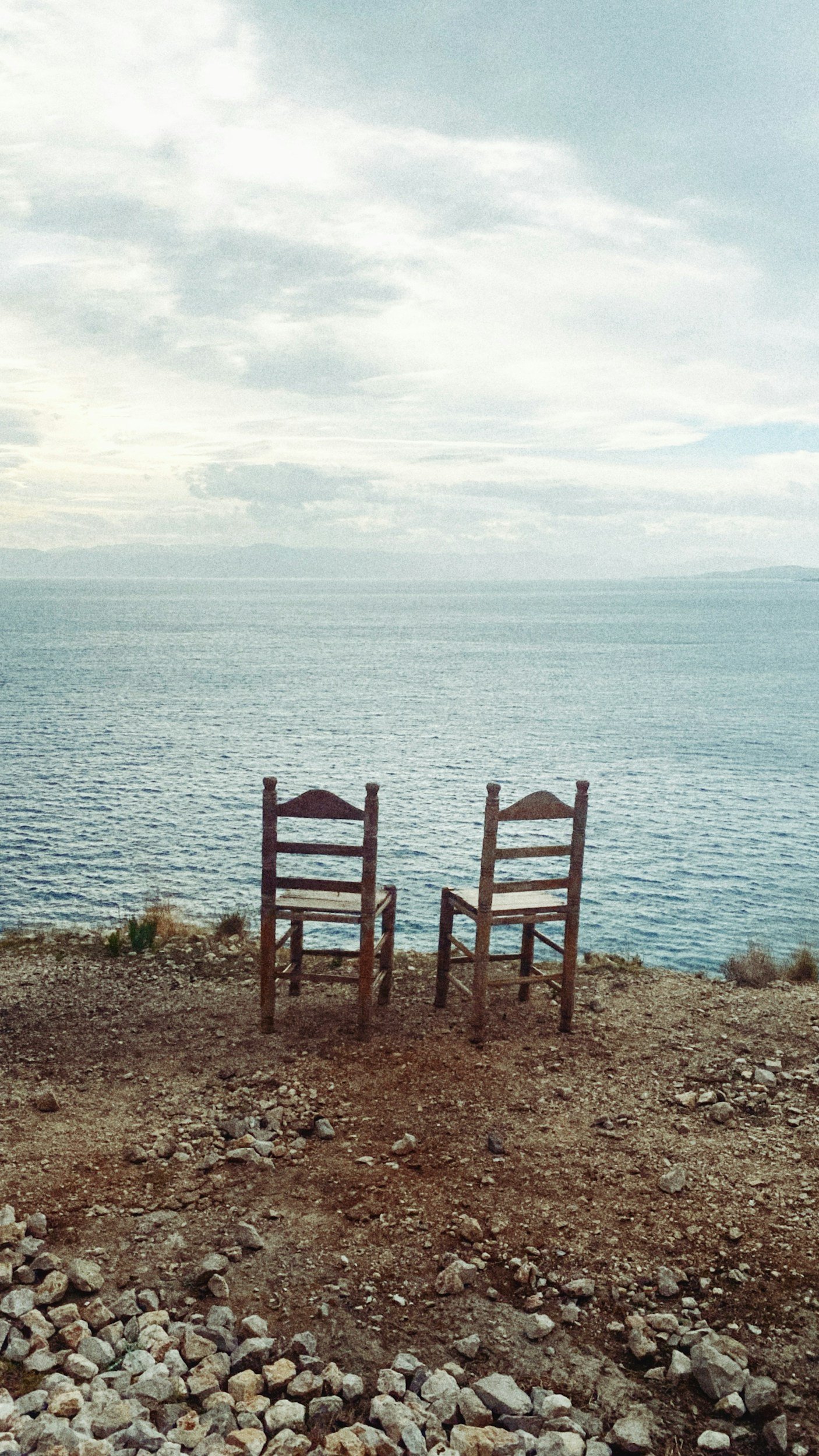 Two wooden chairs facing a large lake or sea under a partly cloudy sky.