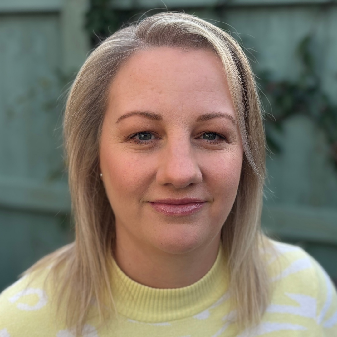 A woman with blonde hair wearing a yellow and white striped top, smiling outdoors with a blurred green background.