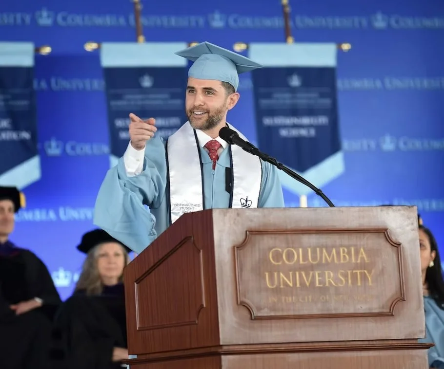 Adam Gerber in a graduation cap and gown speaking at a podium at Columbia University. There are people in graduation robes in the background.