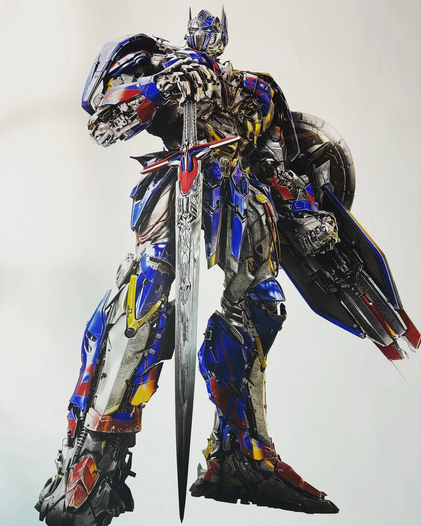 A robot resembling Optimus Prime from Transformers, made from metallic parts and colored in red, blue, yellow, and silver, standing with a large sword and shield.