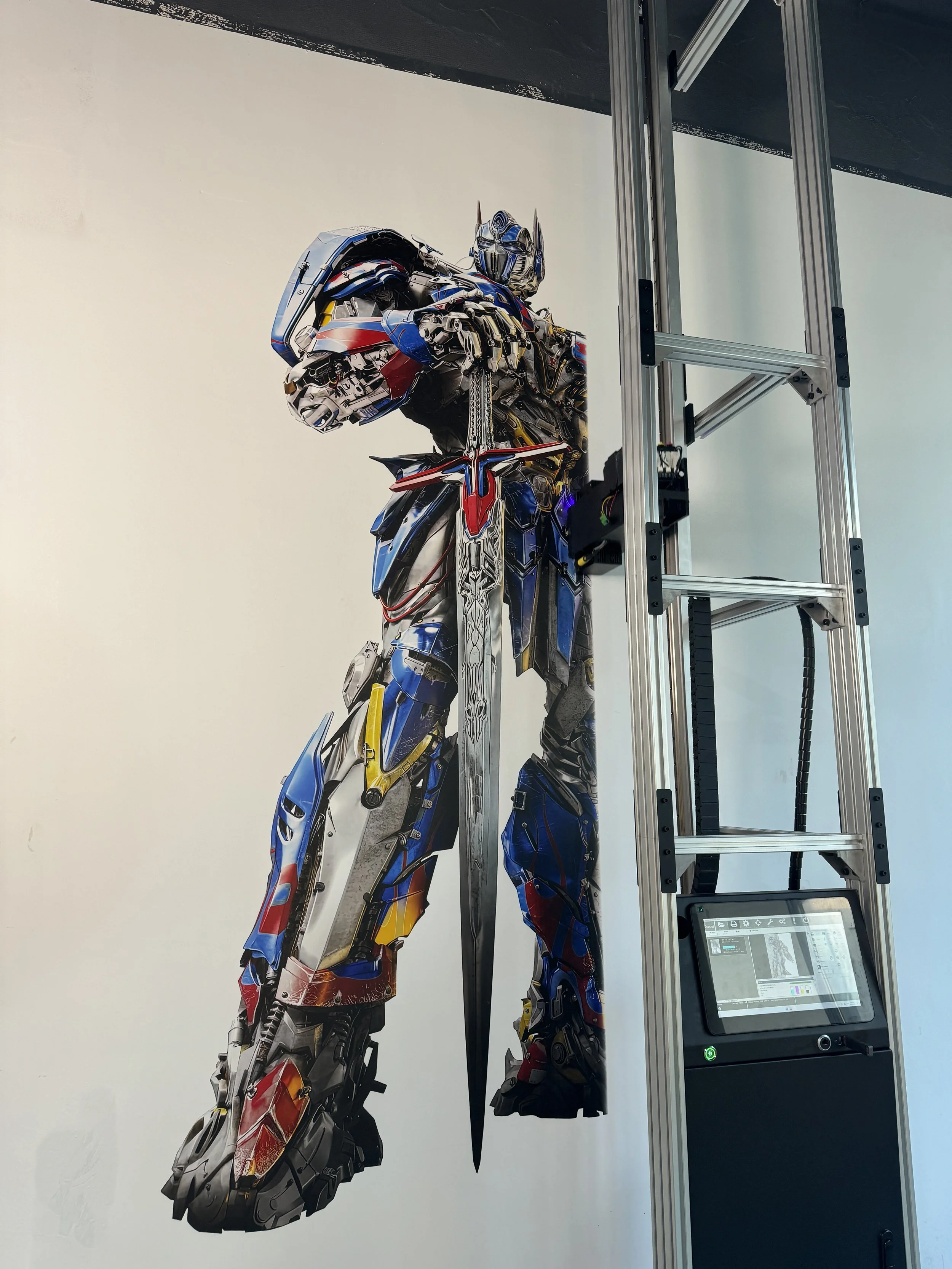 Large depiction of Optimus Prime from Transformers, with robotic metal parts and blue, red, and yellow colors, mounted on a wall beside a ladder with an electronic display.