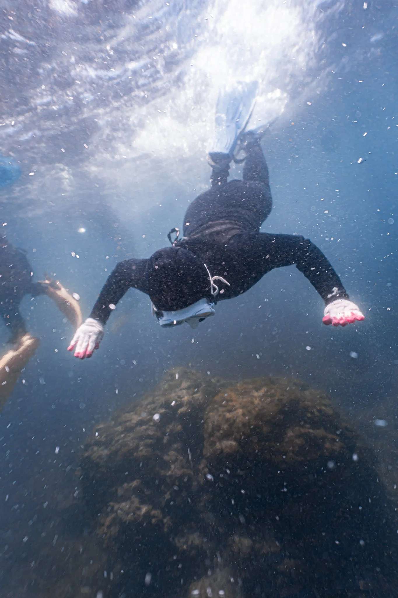 A person in black clothing and pink gloves is swimming underwater, wearing a snorkeling mask and fins, with rocks visible beneath.
