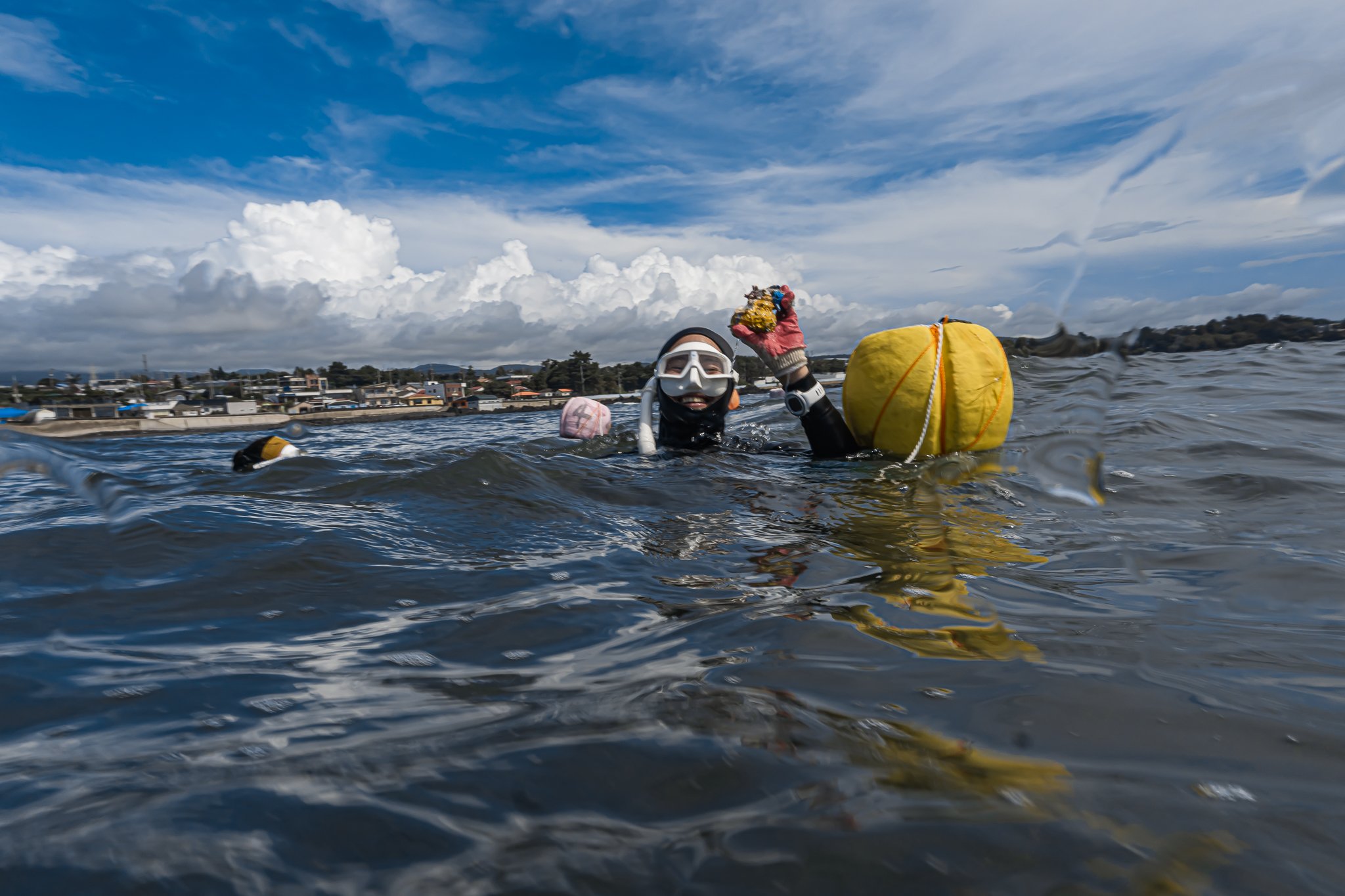 Person in scuba diving gear holding a yellow buoy and coral, with a cloudy sky and shoreline in the background.