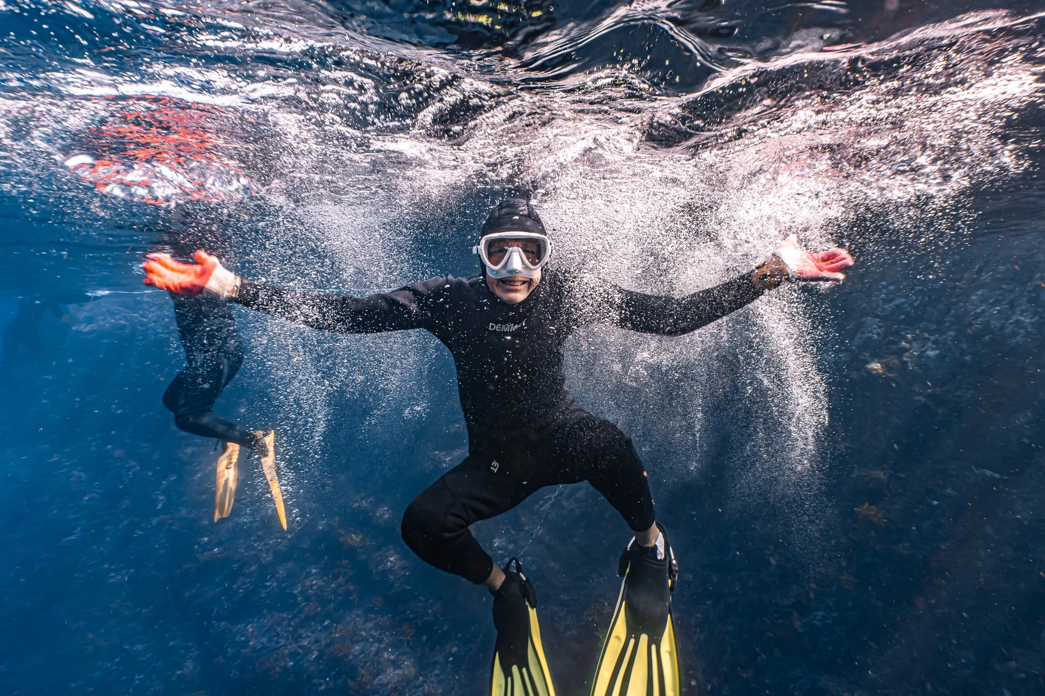 A person wearing snorkeling gear, a black wetsuit, and fins is underwater, smiling and spreading their arms open while surrounded by bubbles.