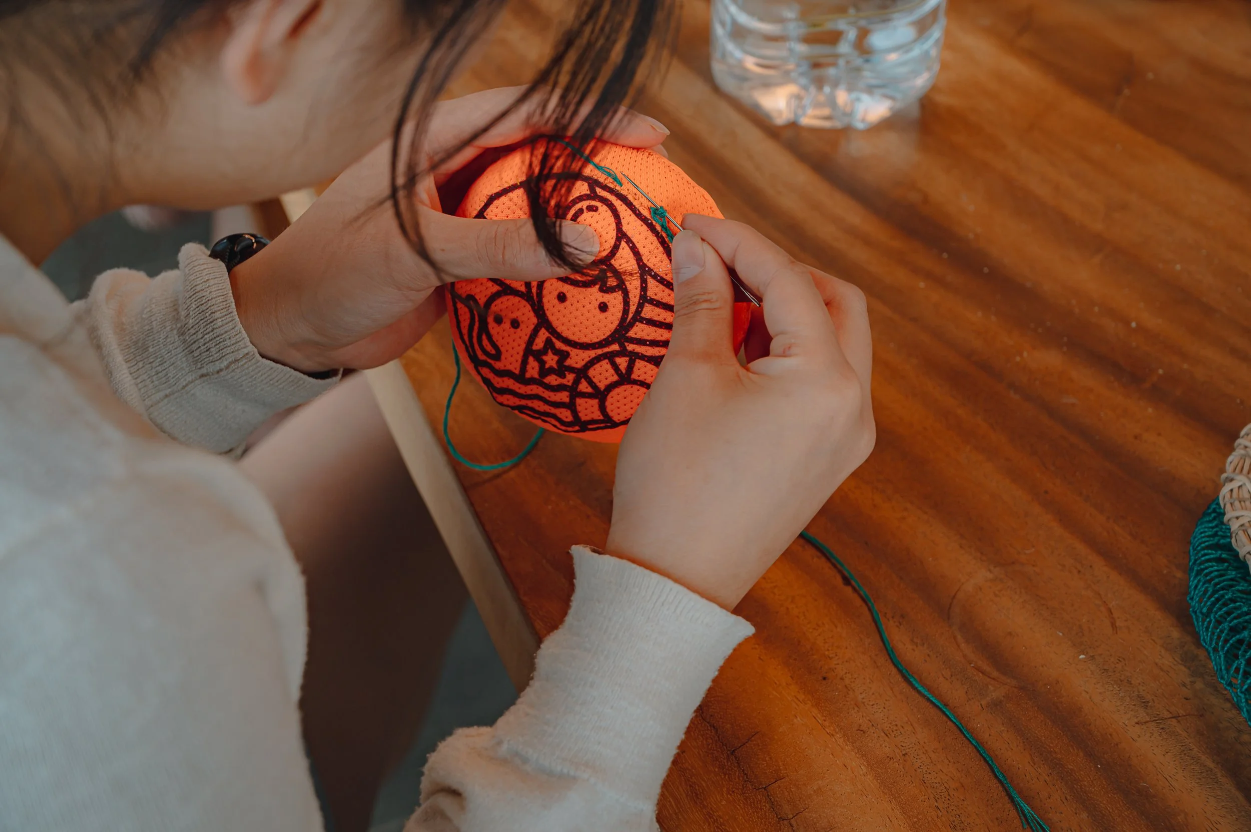 A person threading a needle through a decorative orange lantern with black line art designs of a moon, stars, and a rainbow, on a wooden table with a water bottle and a woven basket nearby.