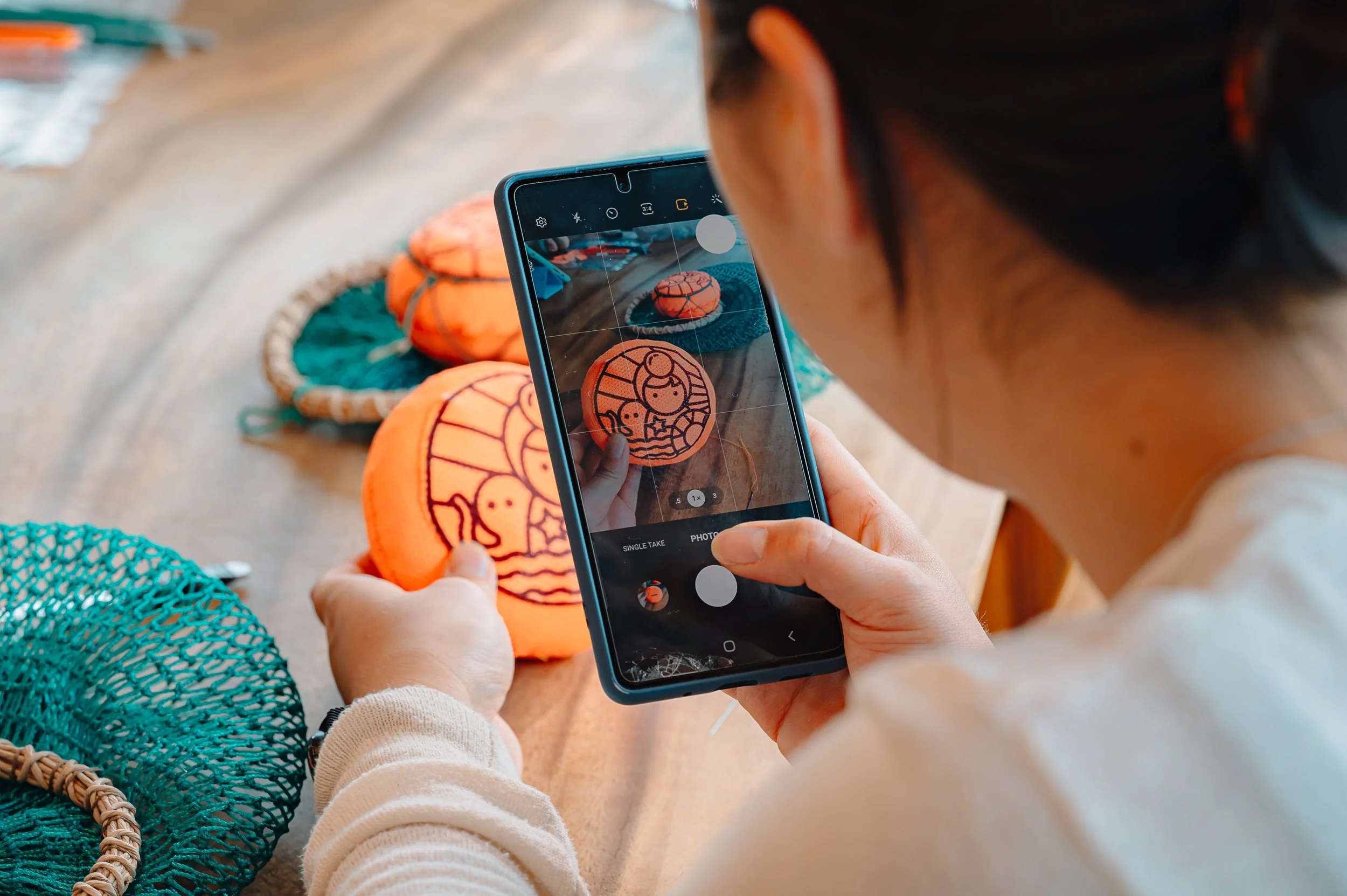 A woman takes a photo of an orange Halloween decoration on a smartphone at a wooden table. The decoration features a cartoon mermaid with seaweed and shells, and there are other similar decorations in the background.