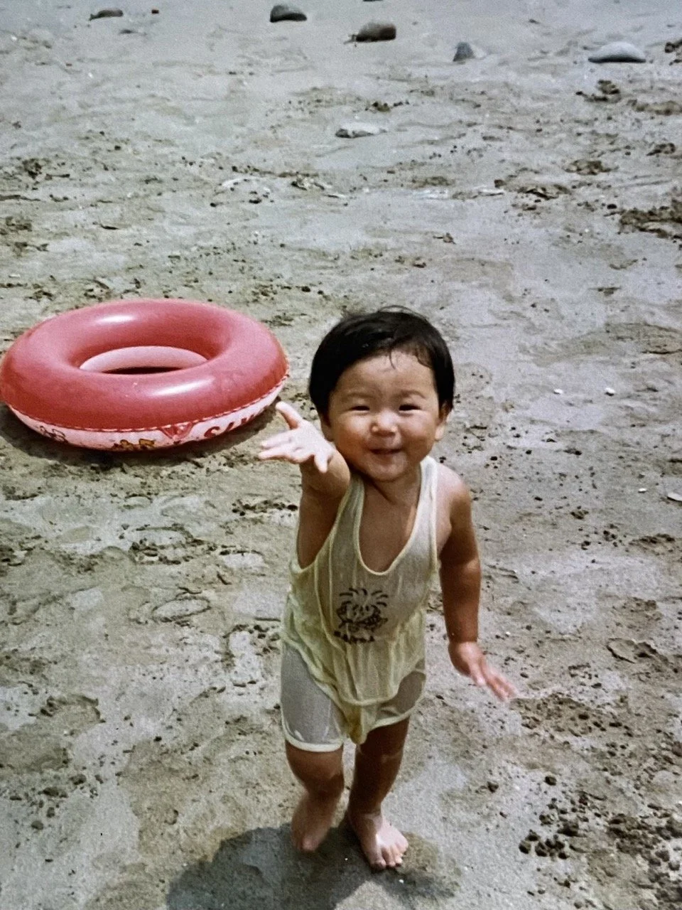Smiling young child on a sandy beach, reaching out with one hand, with a pink inner tube in the background.