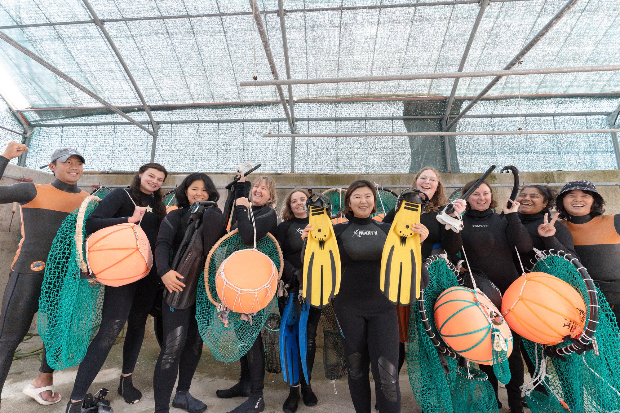 Group of diverse women in wetsuits holding diving gear and orange floats inside a greenhouse or ship hull.