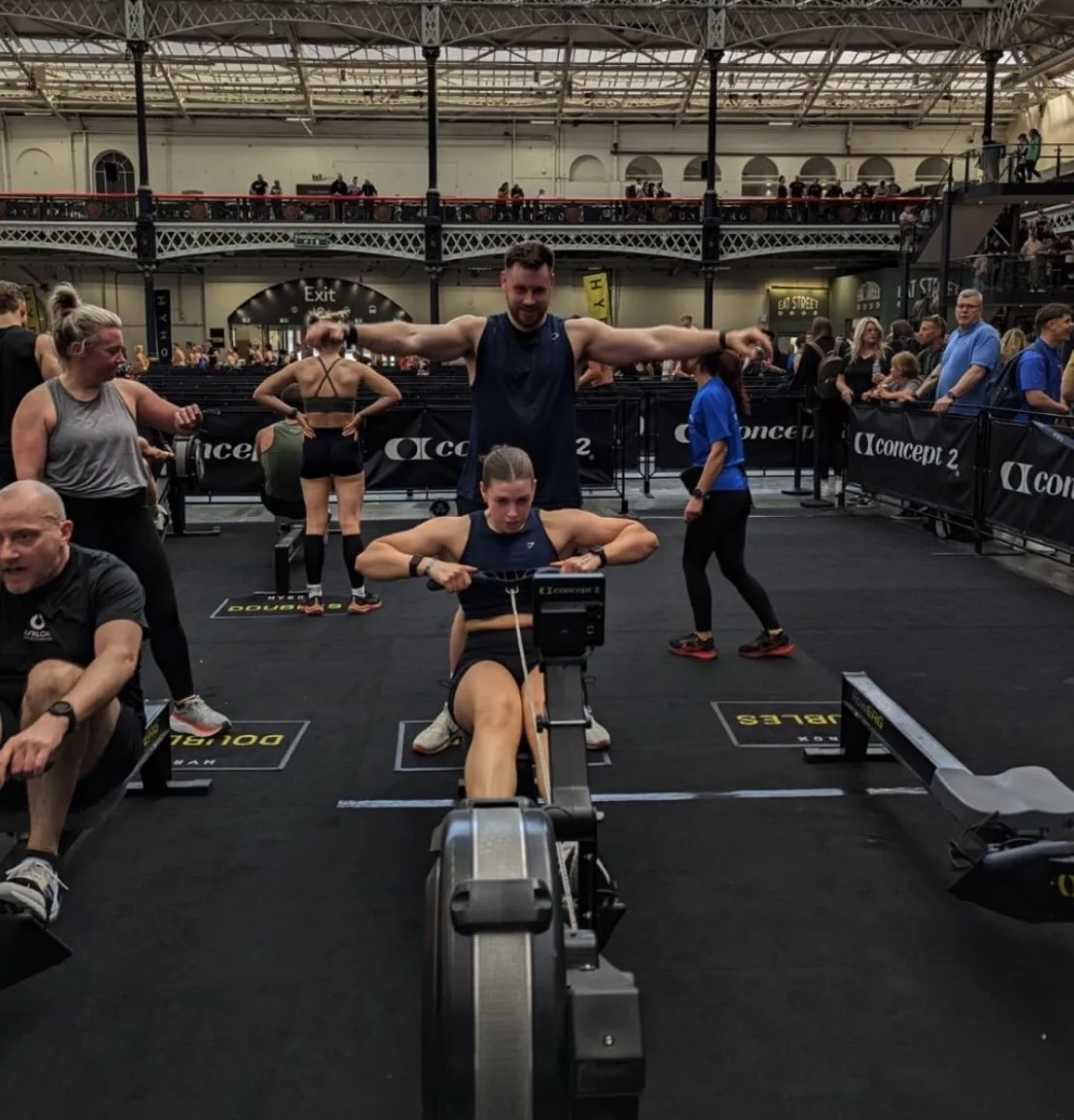 A woman on a rowing machine in a fitness competition, with a muscular coach standing behind her, in a crowded indoor venue with spectators.