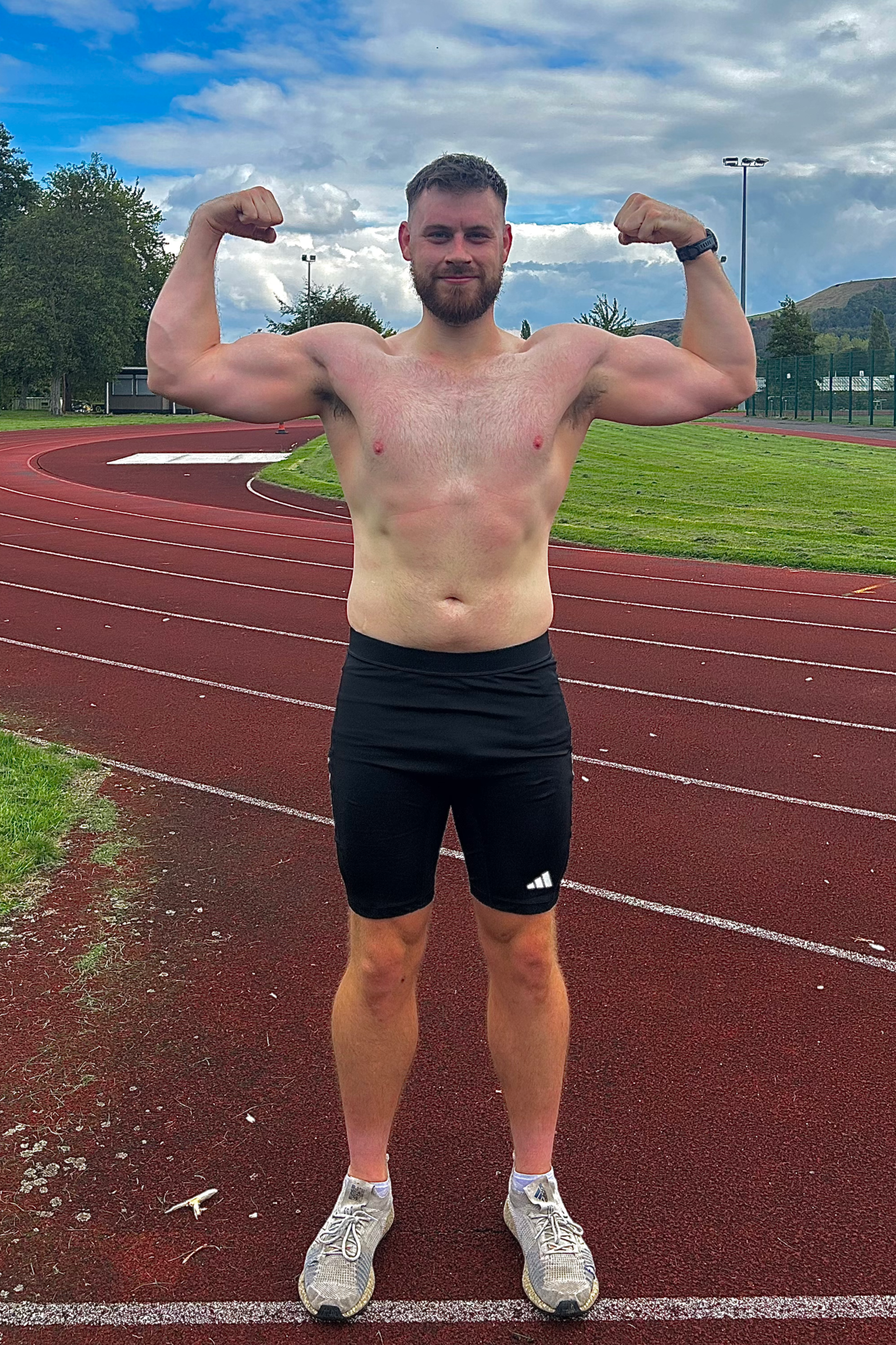 A shirtless man with a beard and short hair poses on a running track, flexing his biceps, wearing black shorts and sneakers, with a cloudy sky and trees in the background.
