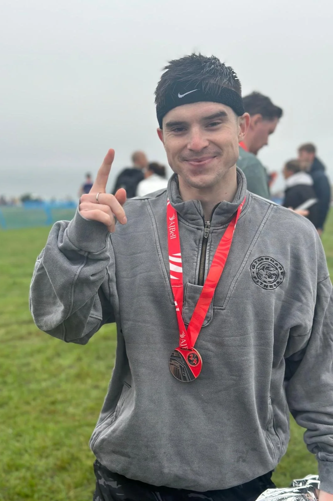 A young man standing outdoors on a grassy field, wearing a gray zip-up jacket, a black headband with a Nike logo, and a red race medal around his neck. He is smiling and pointing upwards with his right index finger. Several people are in the background, some seem to be participating in or spectating at an outdoor sporting event.