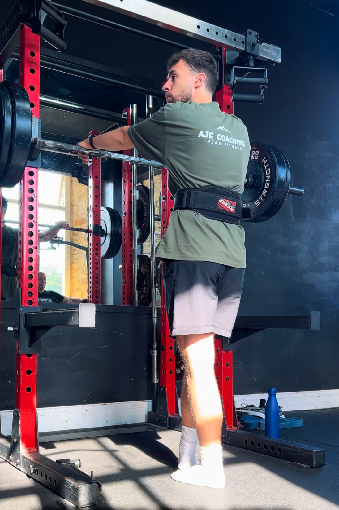 A man in a gray t-shirt and shorts is lifting weights at a gym, standing in front of a red power rack with plates on the barbell. He is using a lifting belt and appears focused on his workout.