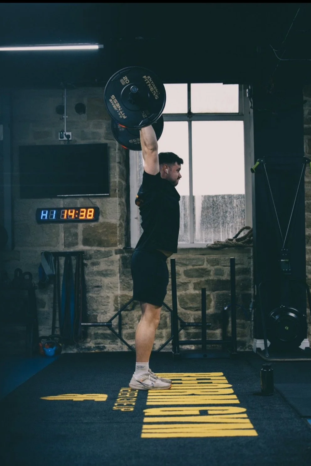 A man lifting a barbell above his head in a gym, with a brick wall and large window in the background.