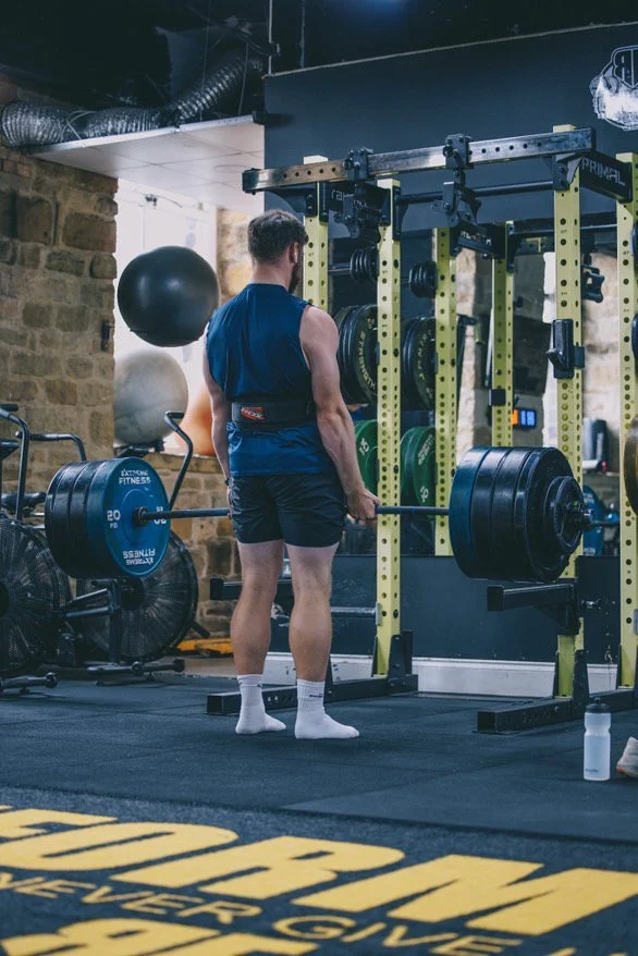 A man lifting a heavy barbell in a gym with black and yellow workout equipment, large medicine balls, and a brick wall in the background.