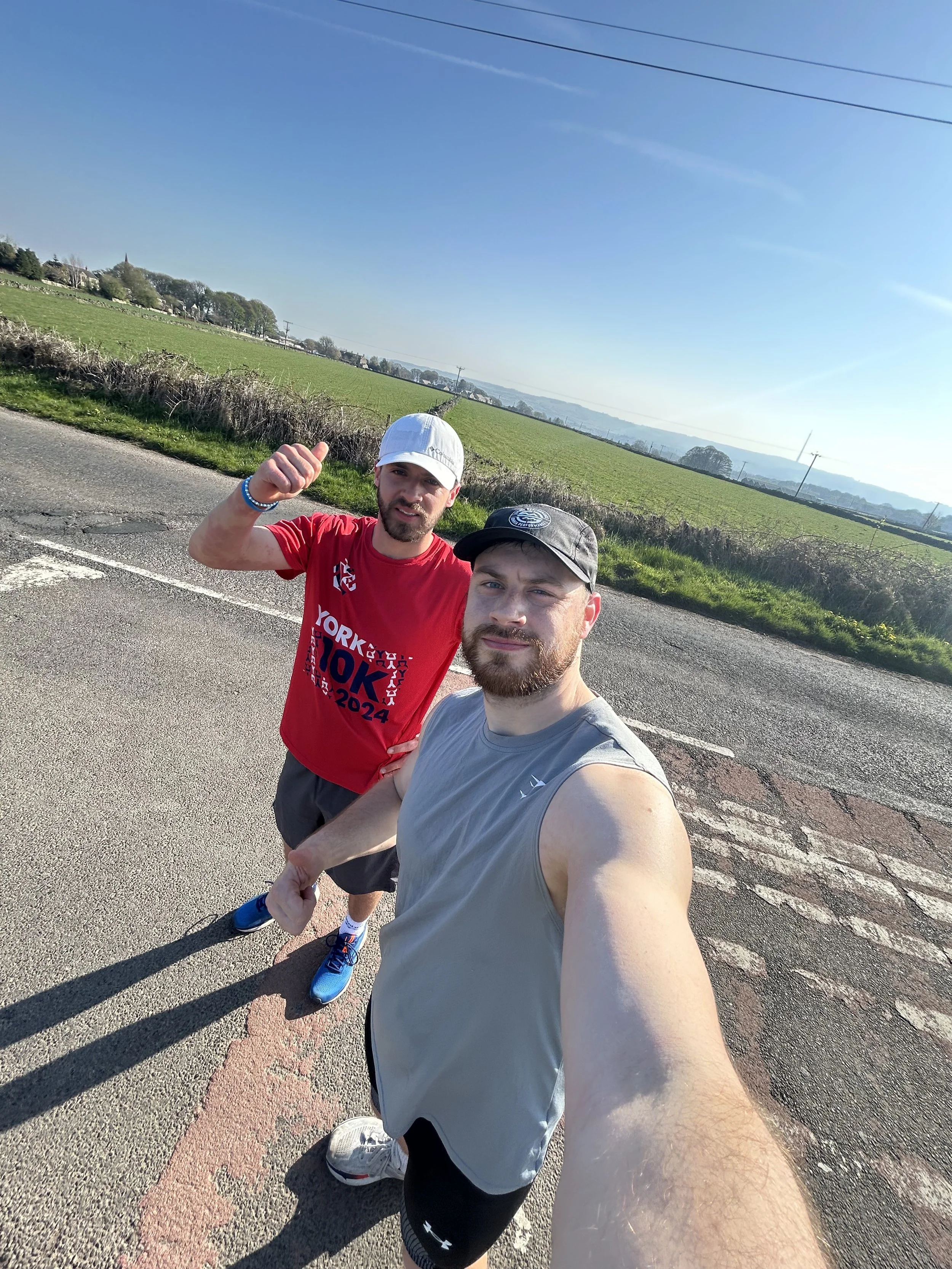 Two men standing on the side of a highway, taking a selfie on a sunny day with green fields and mountains in the background.