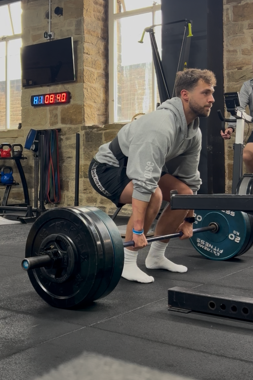A man performing a deadlift exercise with a barbell loaded with weights in a gym. He is wearing a gray hoodie, black shorts, and white socks, and is in a bent-over position lifting the barbell from the ground.
