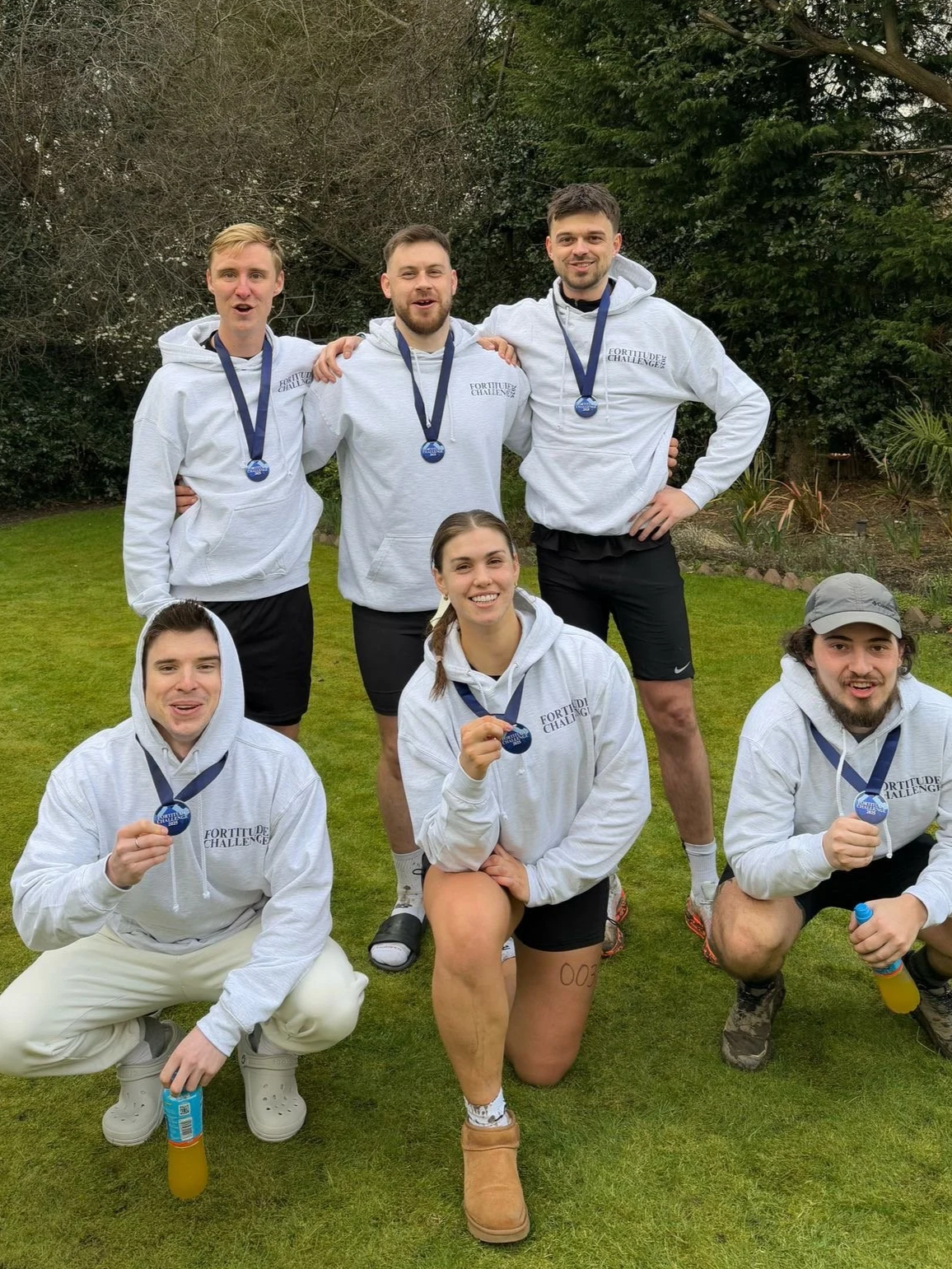 Group of seven young adults outdoors, wearing matching 'Fortitude Challenge' hoodies, displaying medals after participating in a challenge event, with a grassy area and trees in the background.
