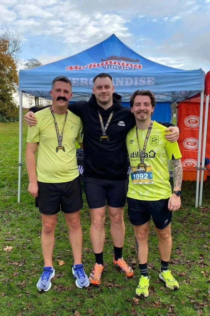 Three men in athletic gear posing together after a race, with medals around their necks and a blue tent in the background.