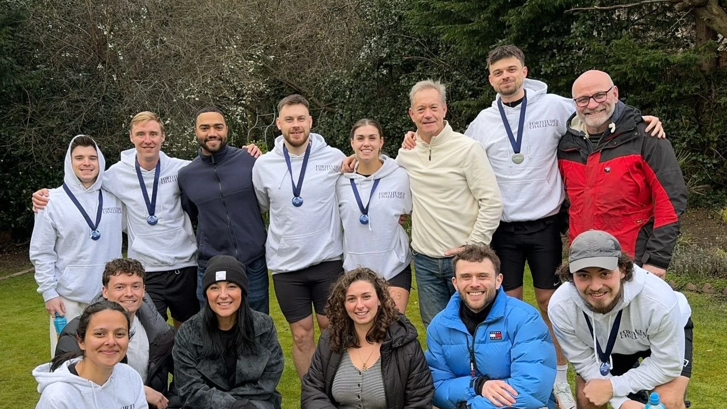 A group of 15 people posing outdoors on grass with trees in the background, some wearing medals and athletic clothing, celebrating after a race or event.