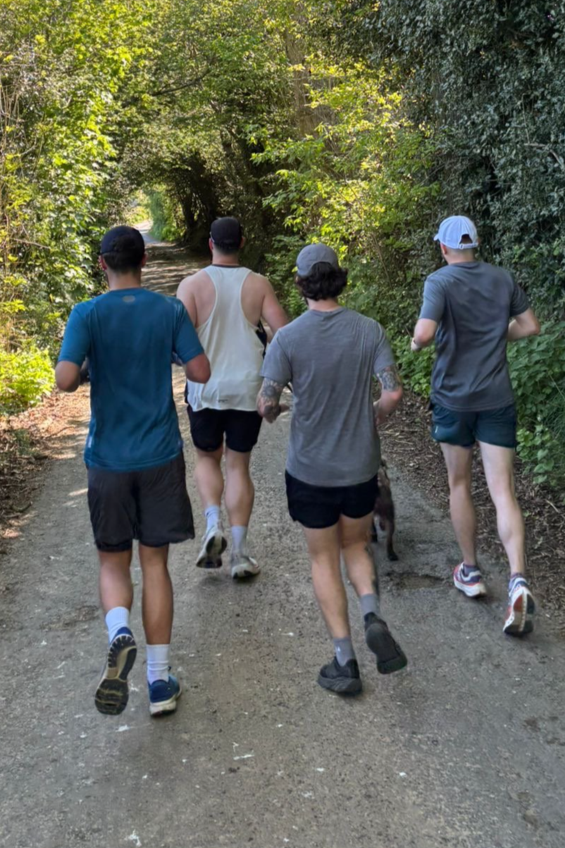 Four young men jogging on a shaded forest trail with trees and bushes on both sides.