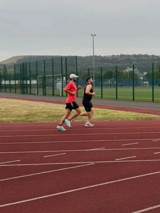 Two people running on an outdoor track, one wearing a red shirt and the other wearing a black tank top, with a fenced sports field and hills in the background.
