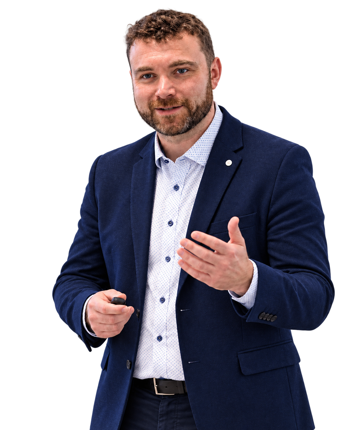 A man with curly hair and a beard, dressed in a dark blue blazer and light shirt, giving a presentation or speech with a remote in his hand and gesturing with his other hand.
