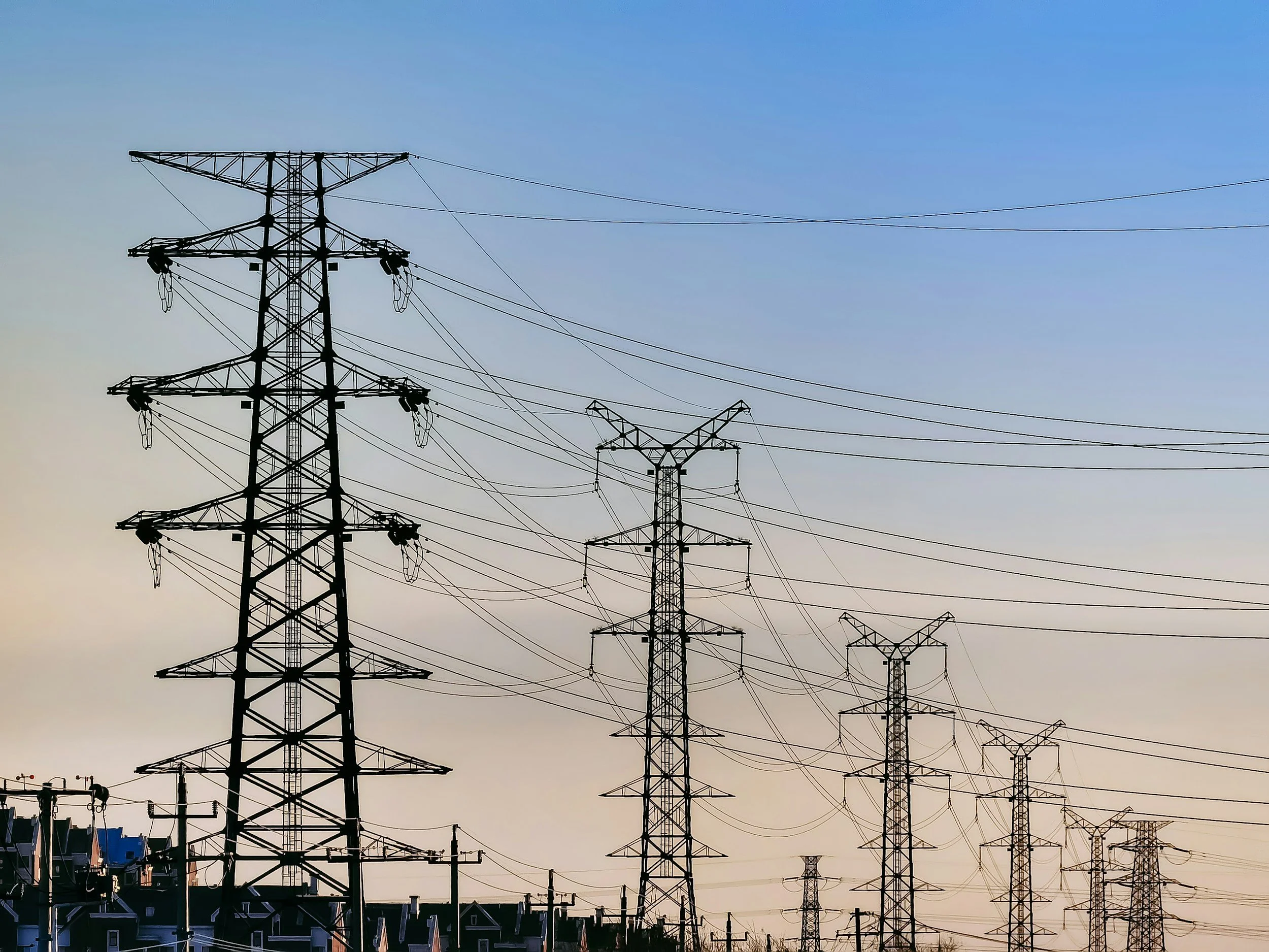 Multiple high-voltage power lines and towers extending into the distance over a residential area during sunset or sunrise.