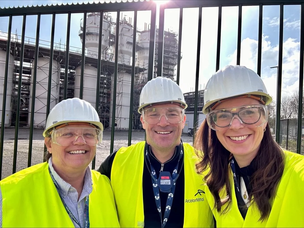 Three workers in high-visibility yellow vests and white safety helmets taking a selfie in front of an industrial facility with large structures and pipes, behind a tall fence.