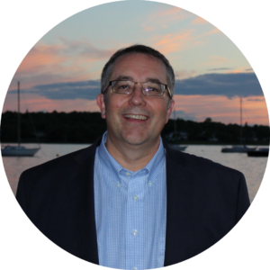 A man in a suit and glasses standing outdoors by a body of water during sunset, smiling.
