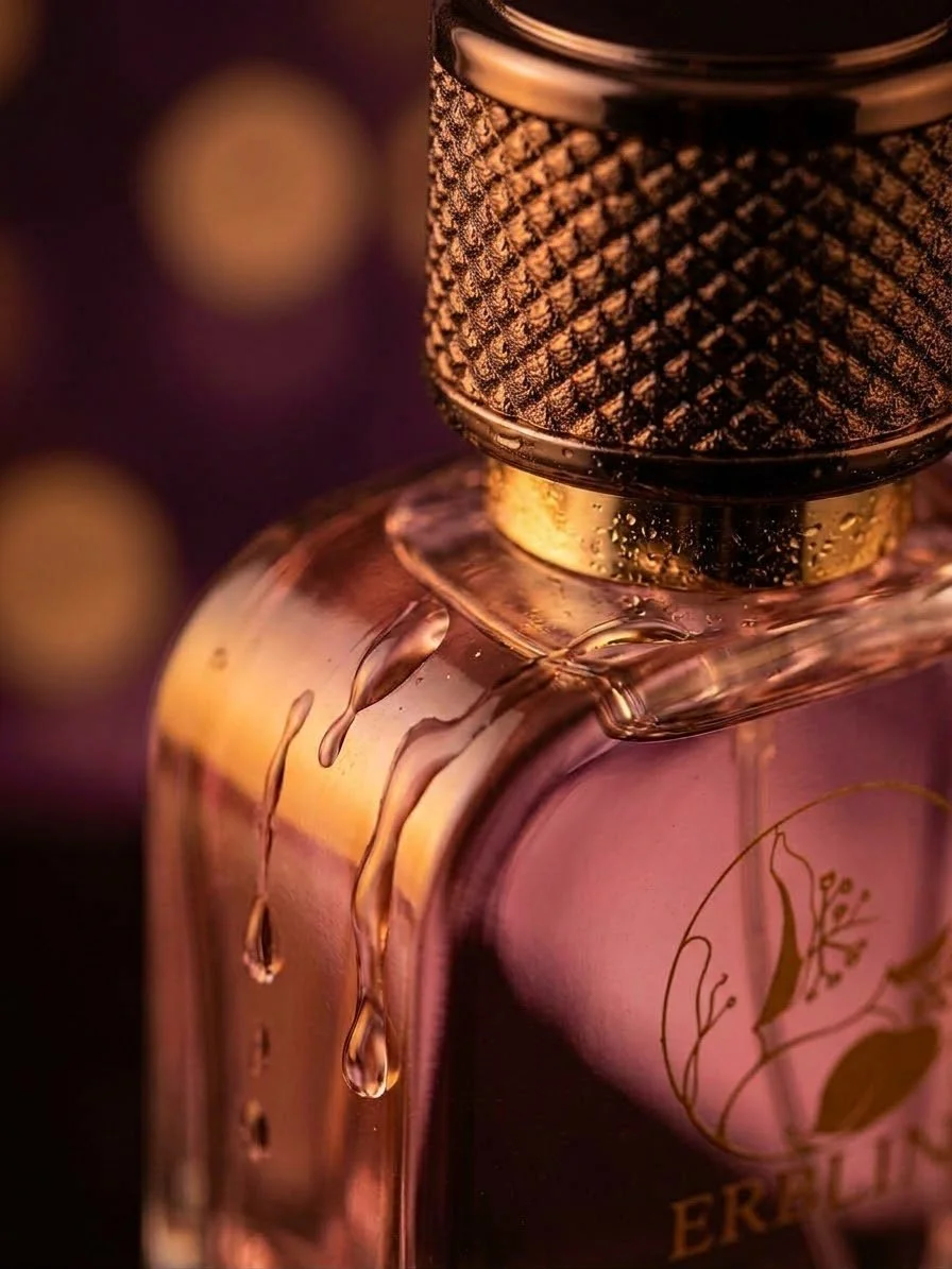 Close-up of a pink perfume bottle with a textured gold and black cap, with liquid droplets on the side, against a blurred background of warm light.