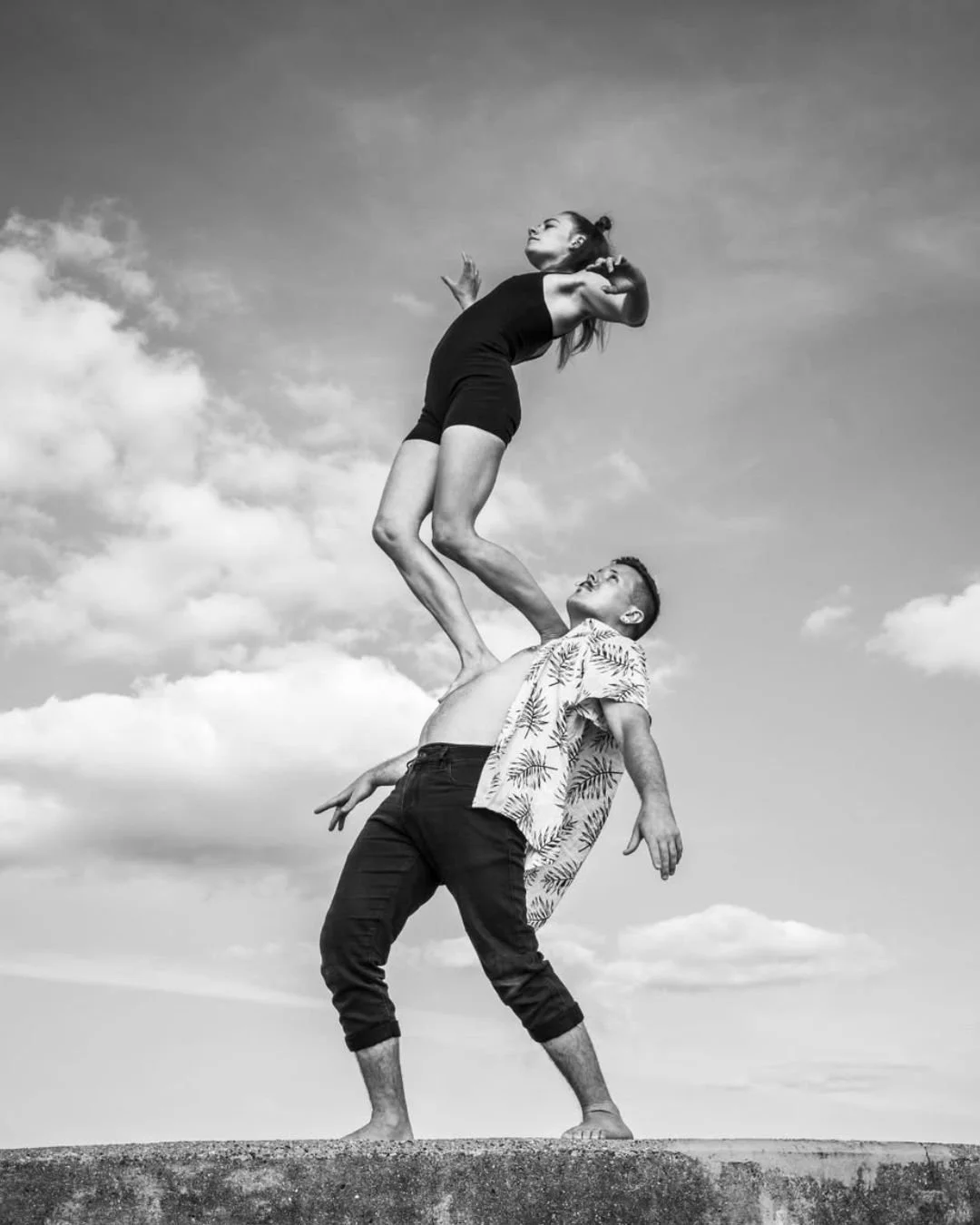 A black-and-white photo of a woman balancing on a man's shoulder as he stands barefoot on a concrete surface, against a cloudy sky.