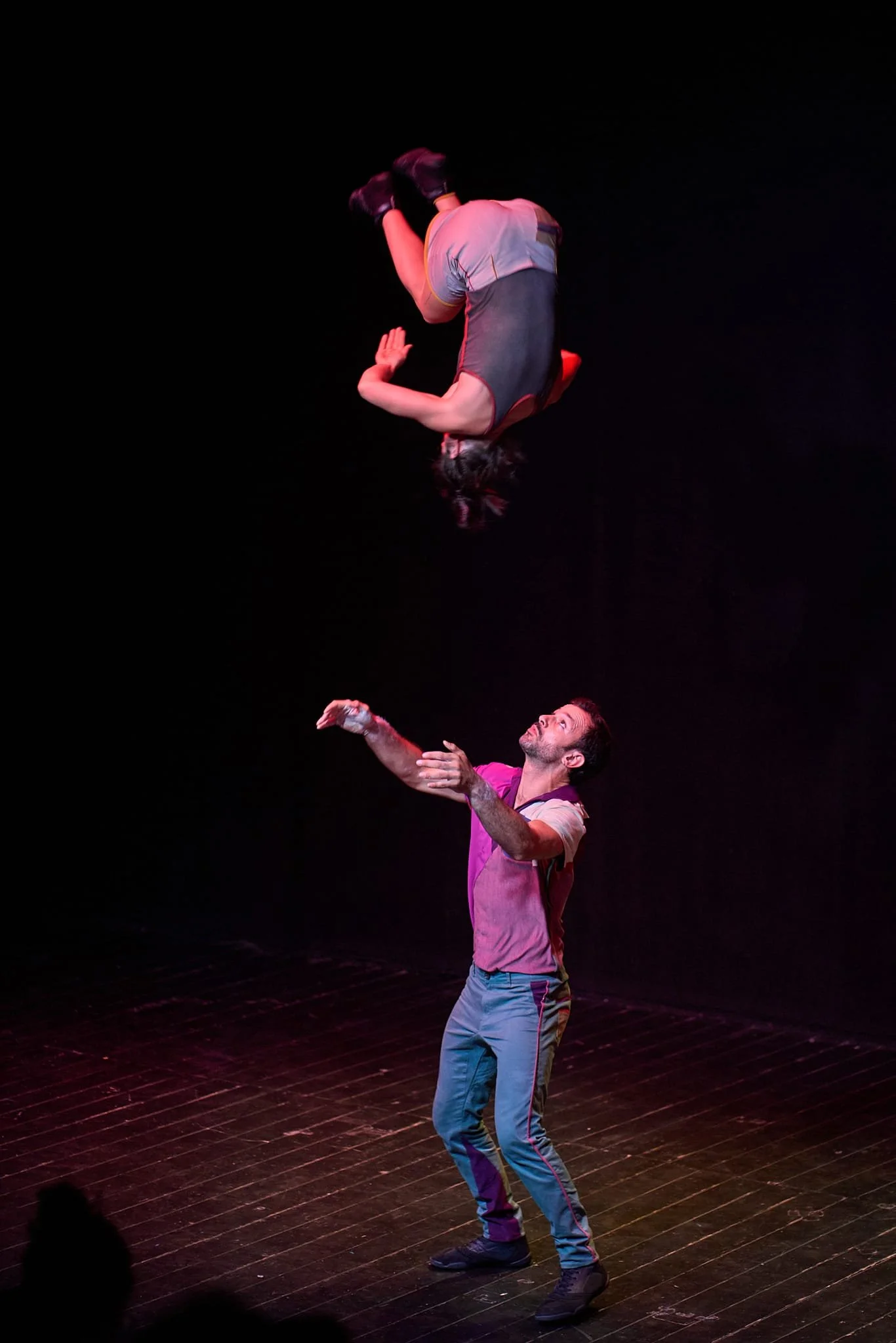 A female performer is captured mid somersault, as a man waits below ready to catch her.