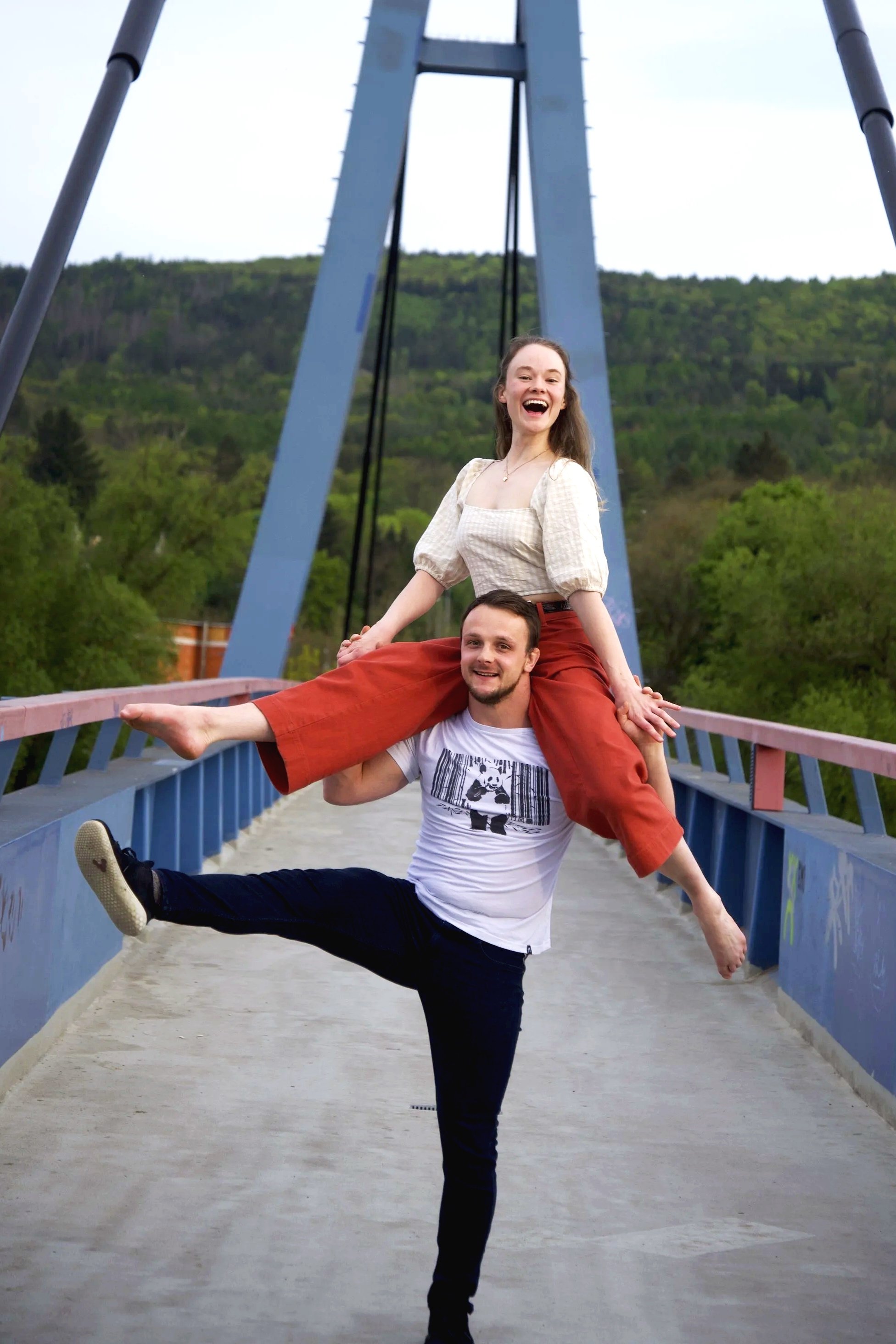 A man and a woman are on a bridge. The woman is sitting on the man's shoulders, and he is holding her ankle to keep her balanced. Both appear to be happy and smiling. The background features green trees and a hillside.
