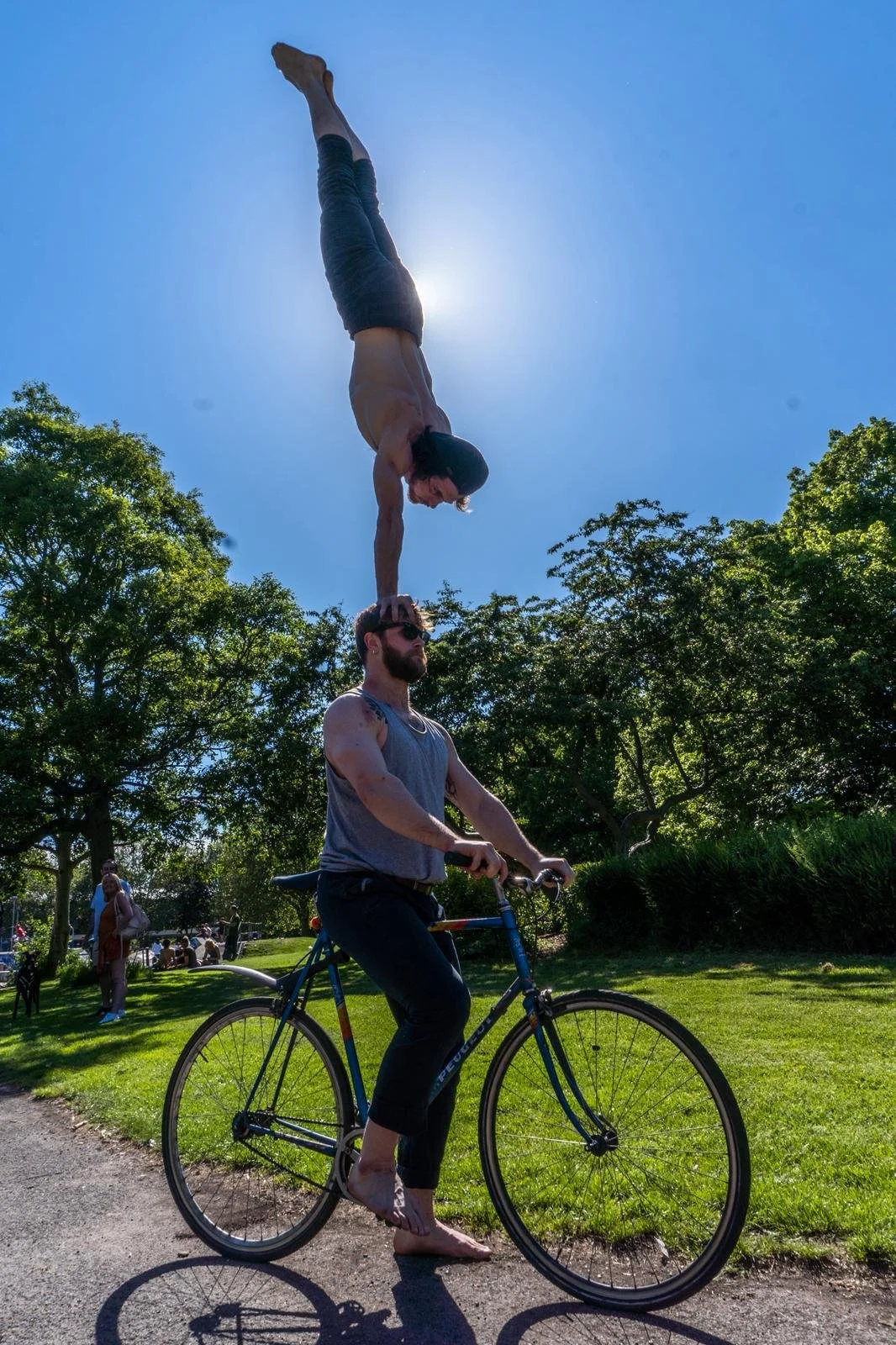 A person riding a bicycle outdoors with a person balancing on their hand in a handstand, positioned above them in the sky with the sun behind.