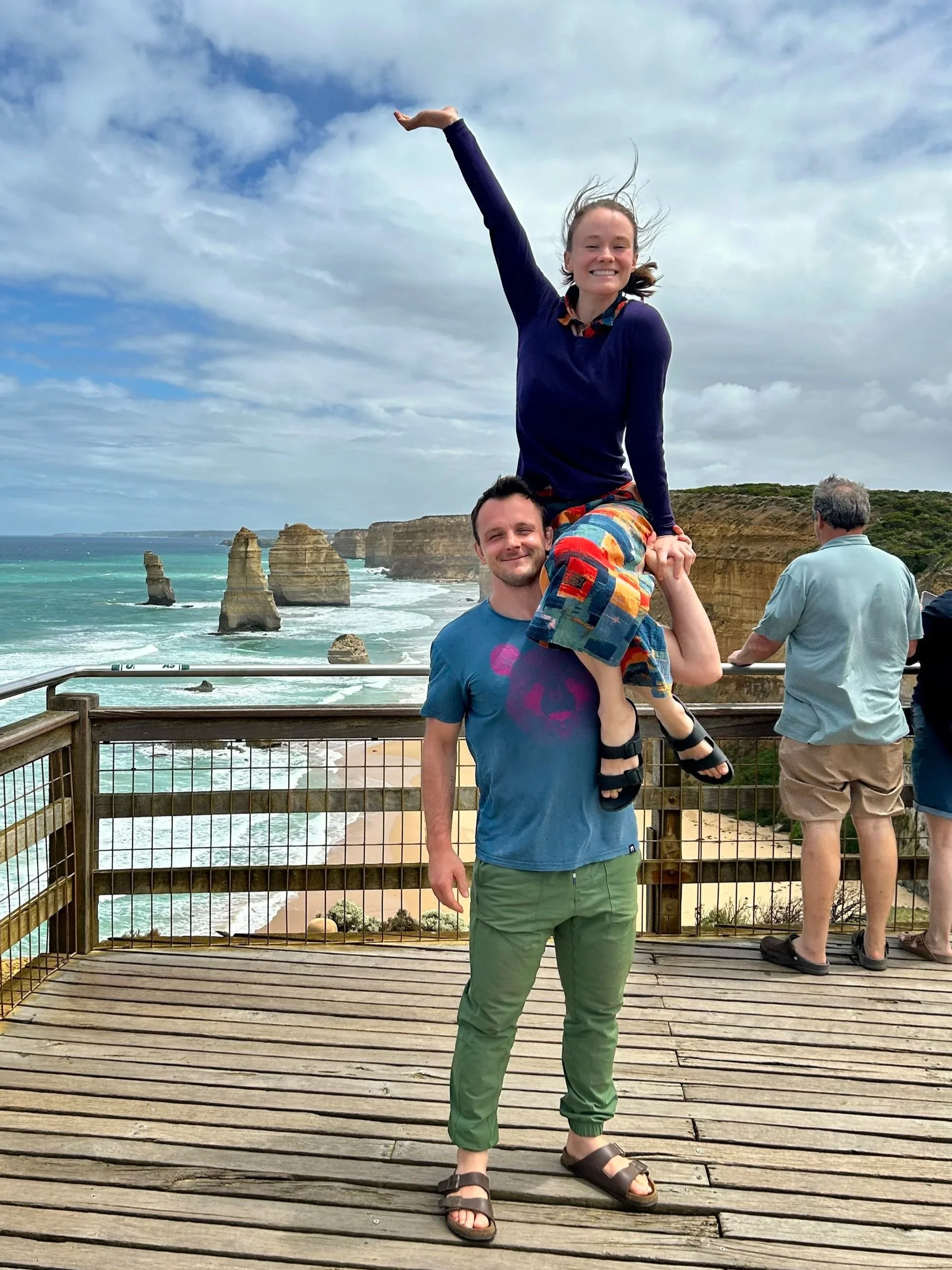 A man is standing on a wooden deck, smiling, with a woman sitting on his shoulders and raising her arm in the air. The background shows the ocean, stone rock formations, and a cloudy sky.