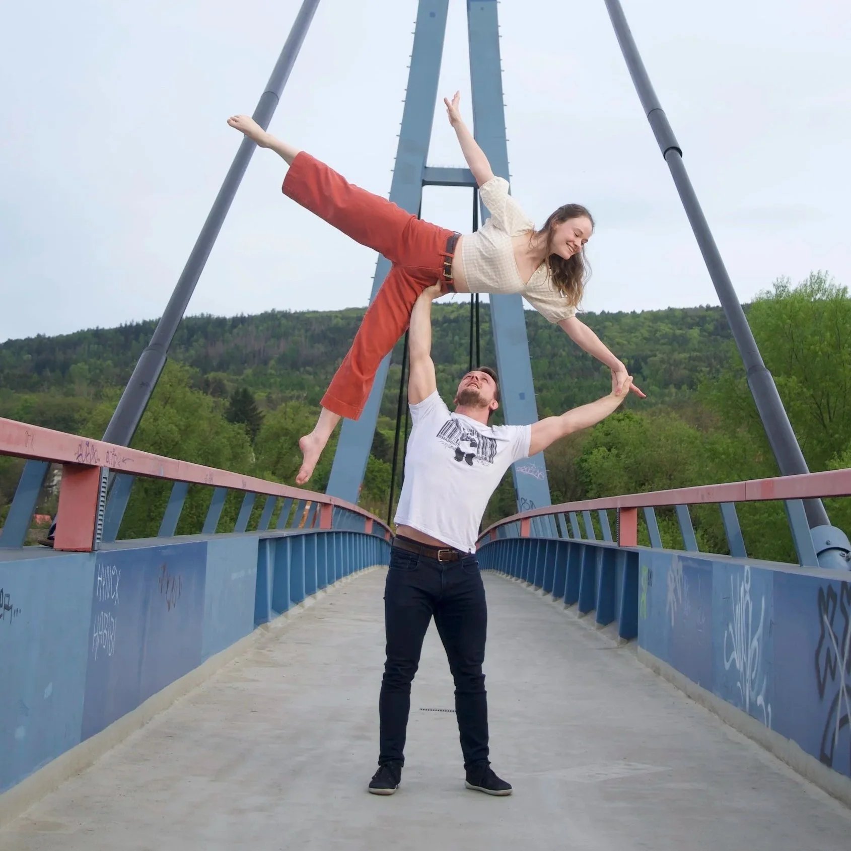 A man lifts a woman in an acrobatic pose above his head on a bridge with a blue metal structure, with green trees and hills in the background.