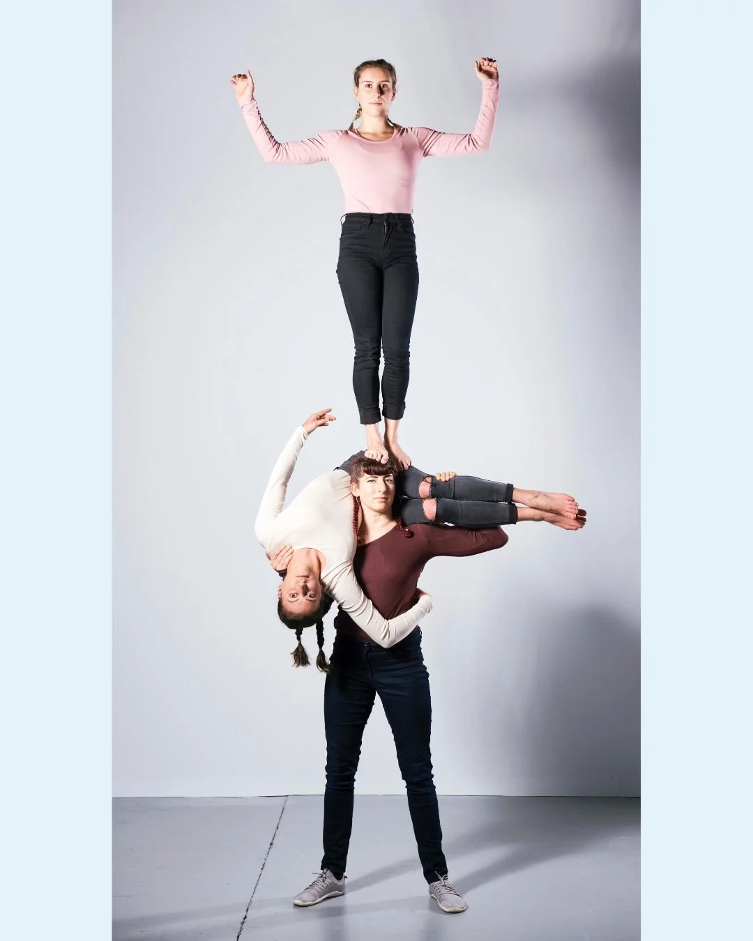 Three women performing a human pyramid. The woman at the bottom supports the other two, with one lying down and the other standing on her shoulders. The woman on top stands with her arms raised.