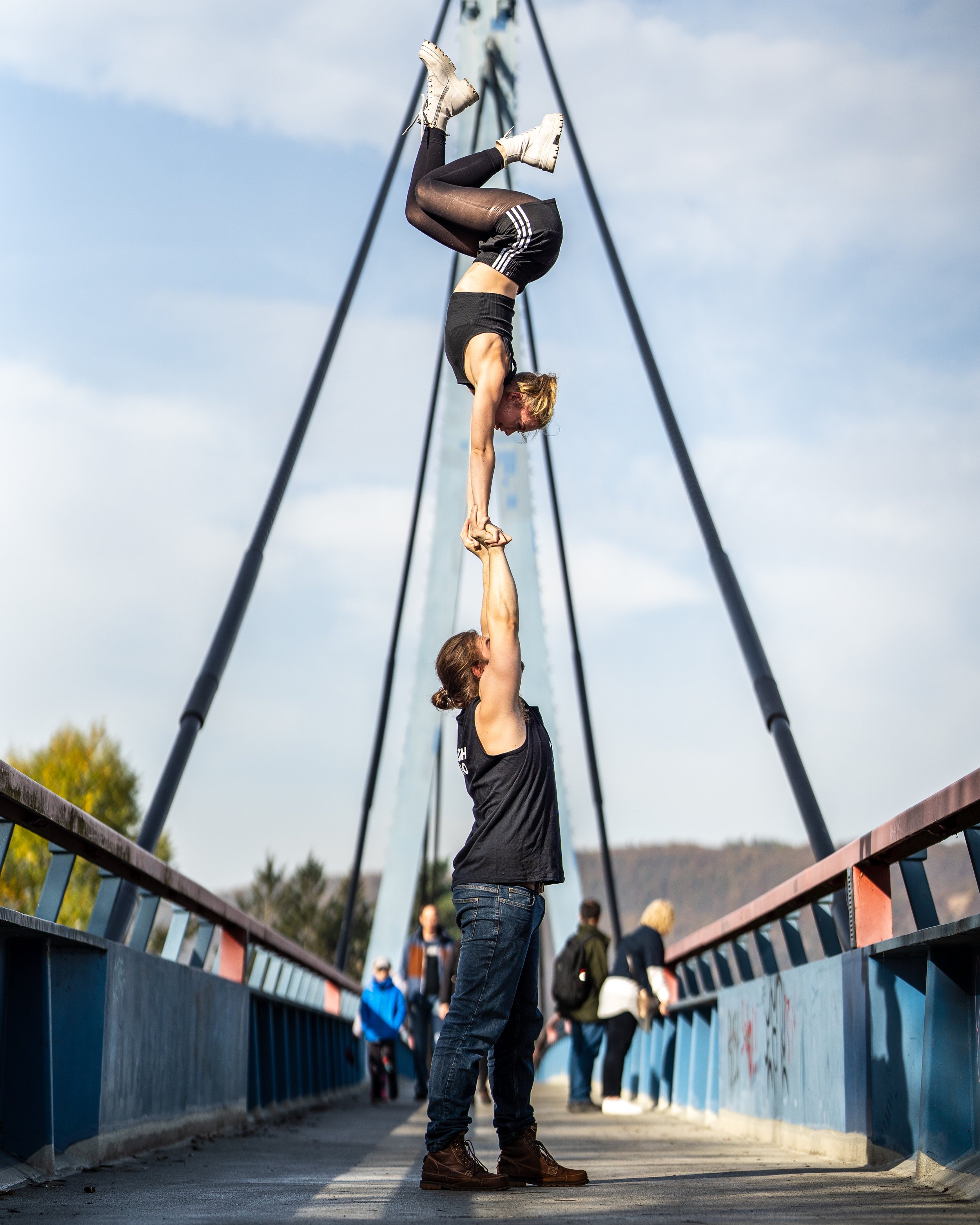 Two women practicing acro yoga on a bridge with a blue railing, with one woman upside down supported by the other.