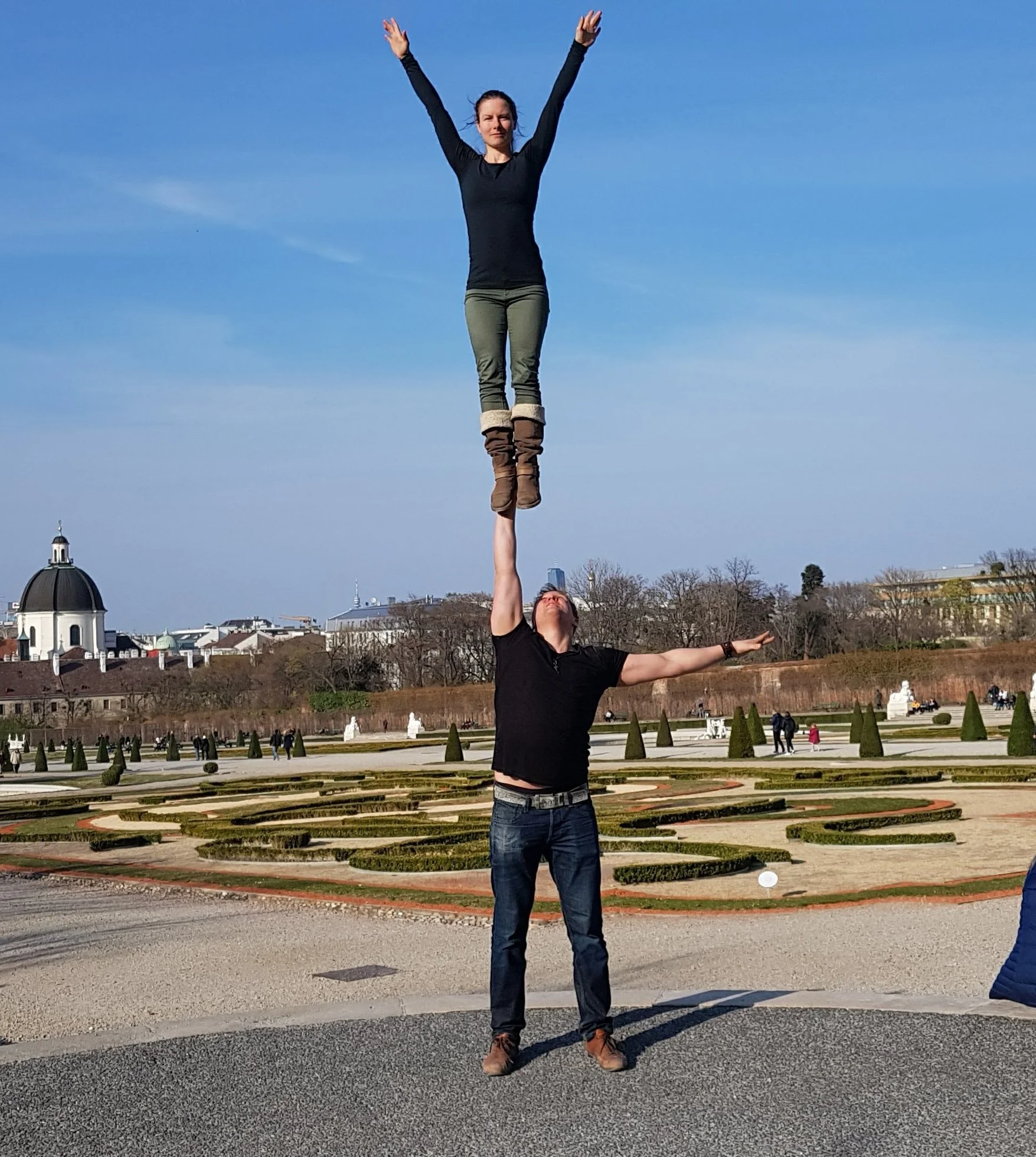 A woman balances on one foot on another person's outstretched hand in an outdoor park with a well-maintained garden, historic buildings, and a clear blue sky in the background.