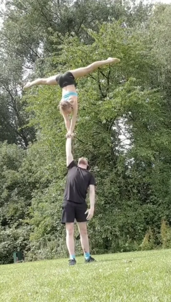 A person standing on grass holding a girl upside down by her hand. The girl is balanced with her body extended horizontally, performing a handstand on the man's hand. There are trees in the background.