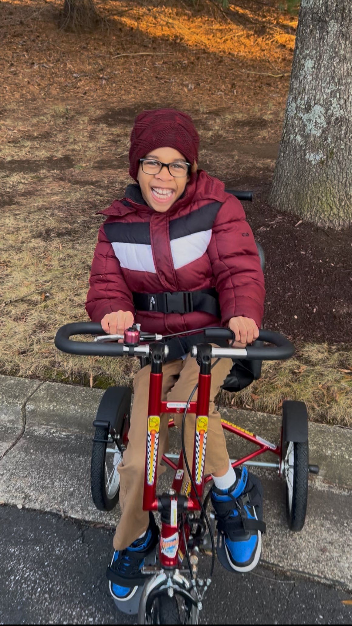 A young boy smiling excitedly while sitting on a red adaptive tricycle outdoors next to a tree.