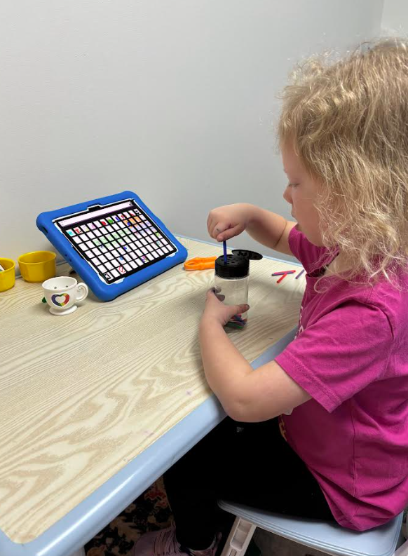 A young girl with curly blonde hair wearing a pink t-shirt is pouring small colorful items from a container into a jar, by a table with a tablet, small cups, and tools.