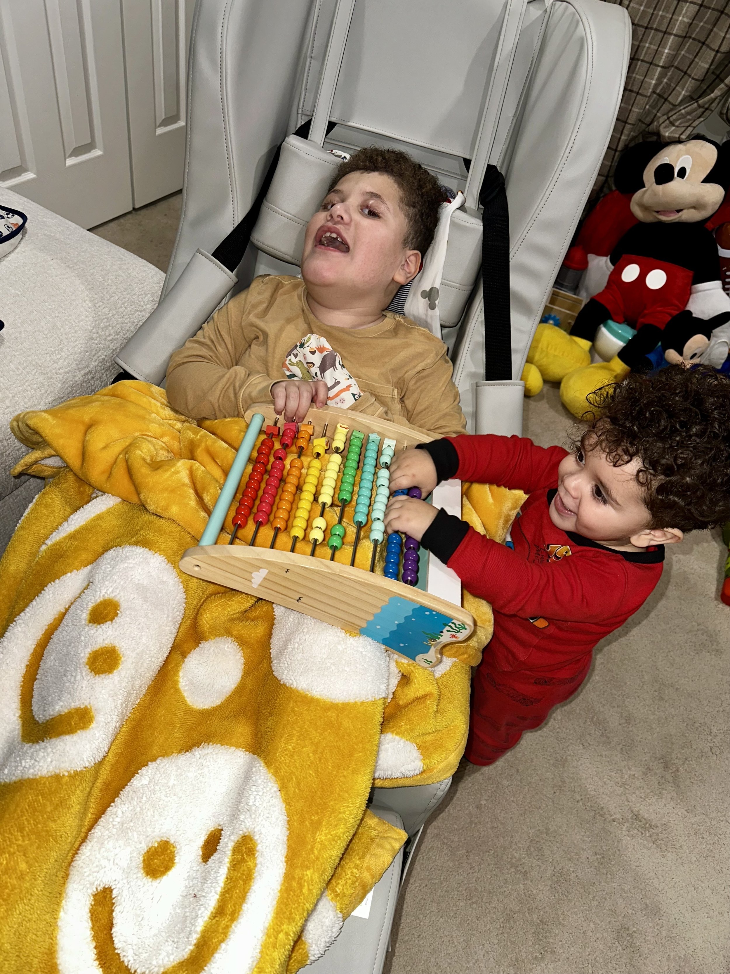 A young boy in a beige shirt sitting in a specialty wheelchair, and a young girl with curly hair in red pajamas playing with a colorful abacus on a yellow smiley face blanket, in a room with Disney plush toys in the background.