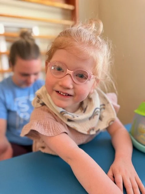 A young girl with glasses smiling at the camera, sitting at a table with another girl in the background.