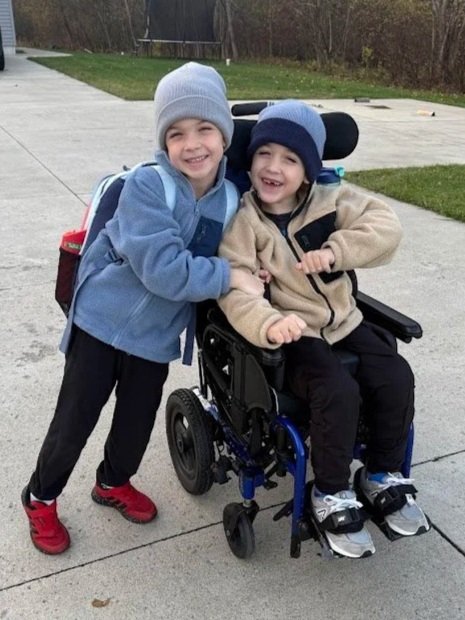 Two young boys smiling, one standing and the other in a wheelchair outdoors on a paved path, with trees and grass in the background.