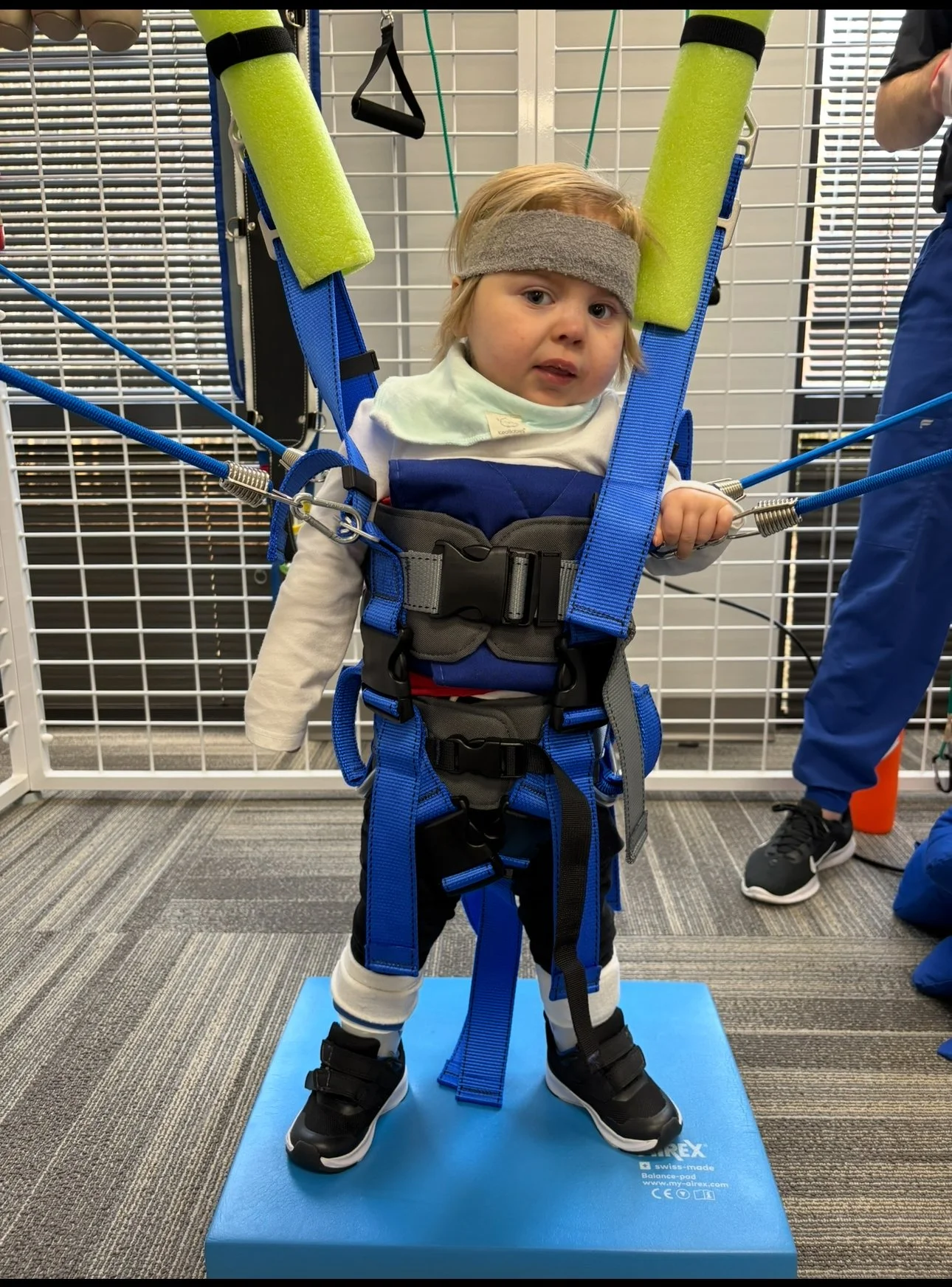 Young child with blond hair wearing a gray headband and a white long-sleeve shirt, standing on a blue balance pad while secured in a harness with blue straps and foam rollers on the harness. The child is holding a handle with their right hand. The scene appears to be in a medical or therapy setting, with a white grid wall behind and an adult partially visible on the right side.