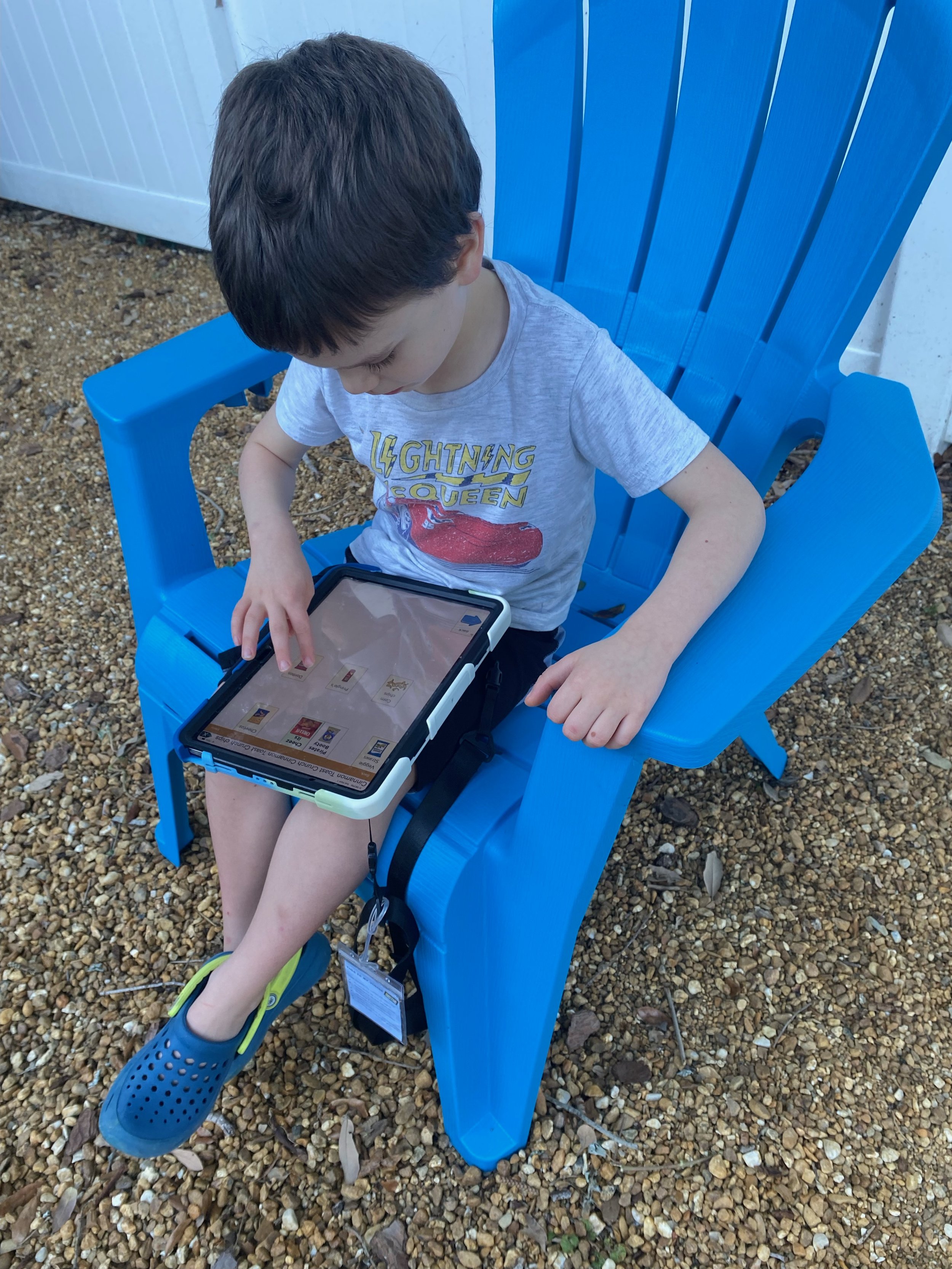 Young boy sitting on a bright blue outdoor bench, using a tablet device, wearing a Lightening Queen t-shirt, shorts, and blue crocs, on gravel ground.